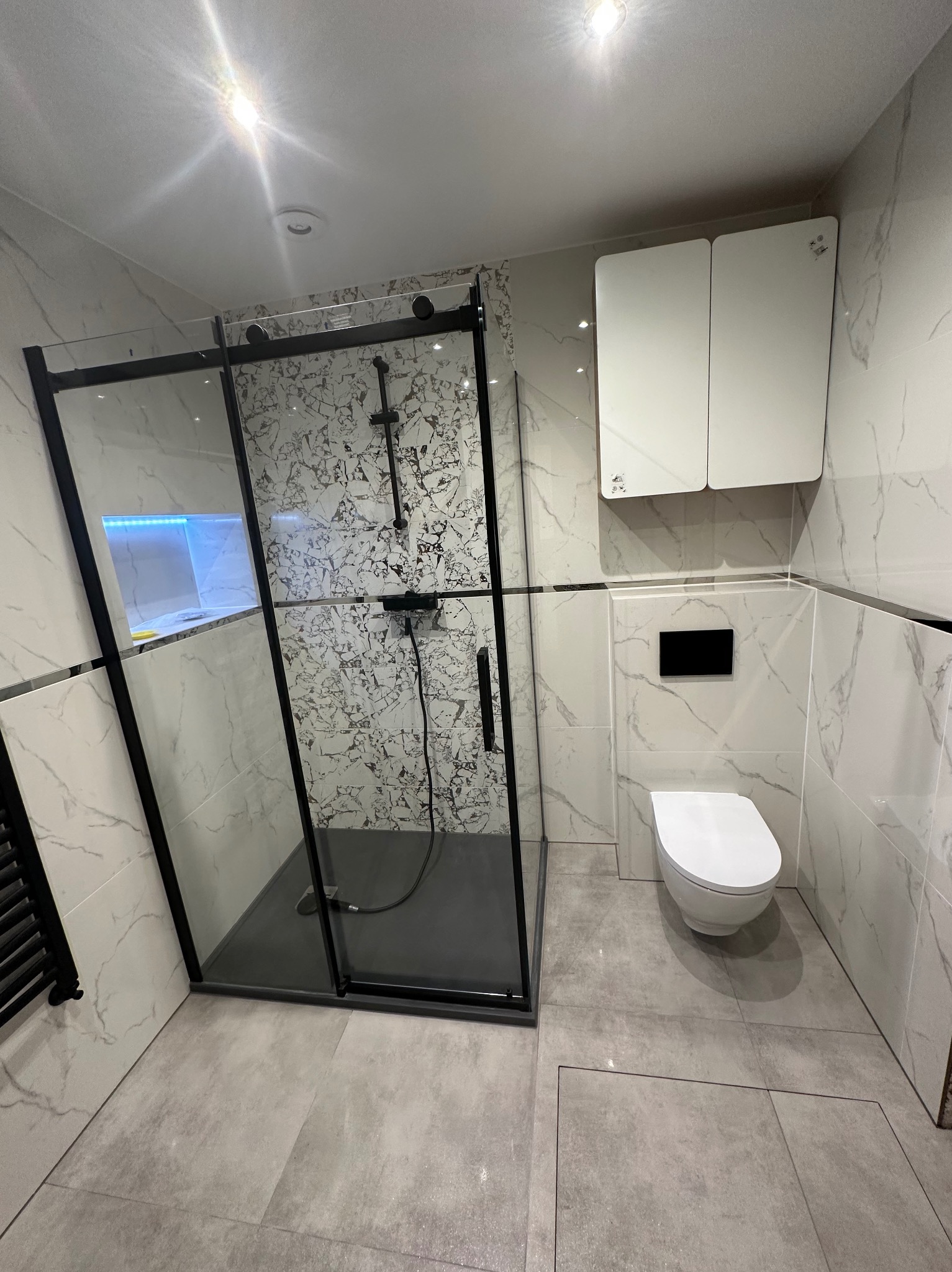 Nowoczesna łazienka z kabiną prysznicową typu walk-in z czarną ramą, marmurowe płytki na ścianach, szara podłoga, podwieszana toaleta i białe szafki wiszące.