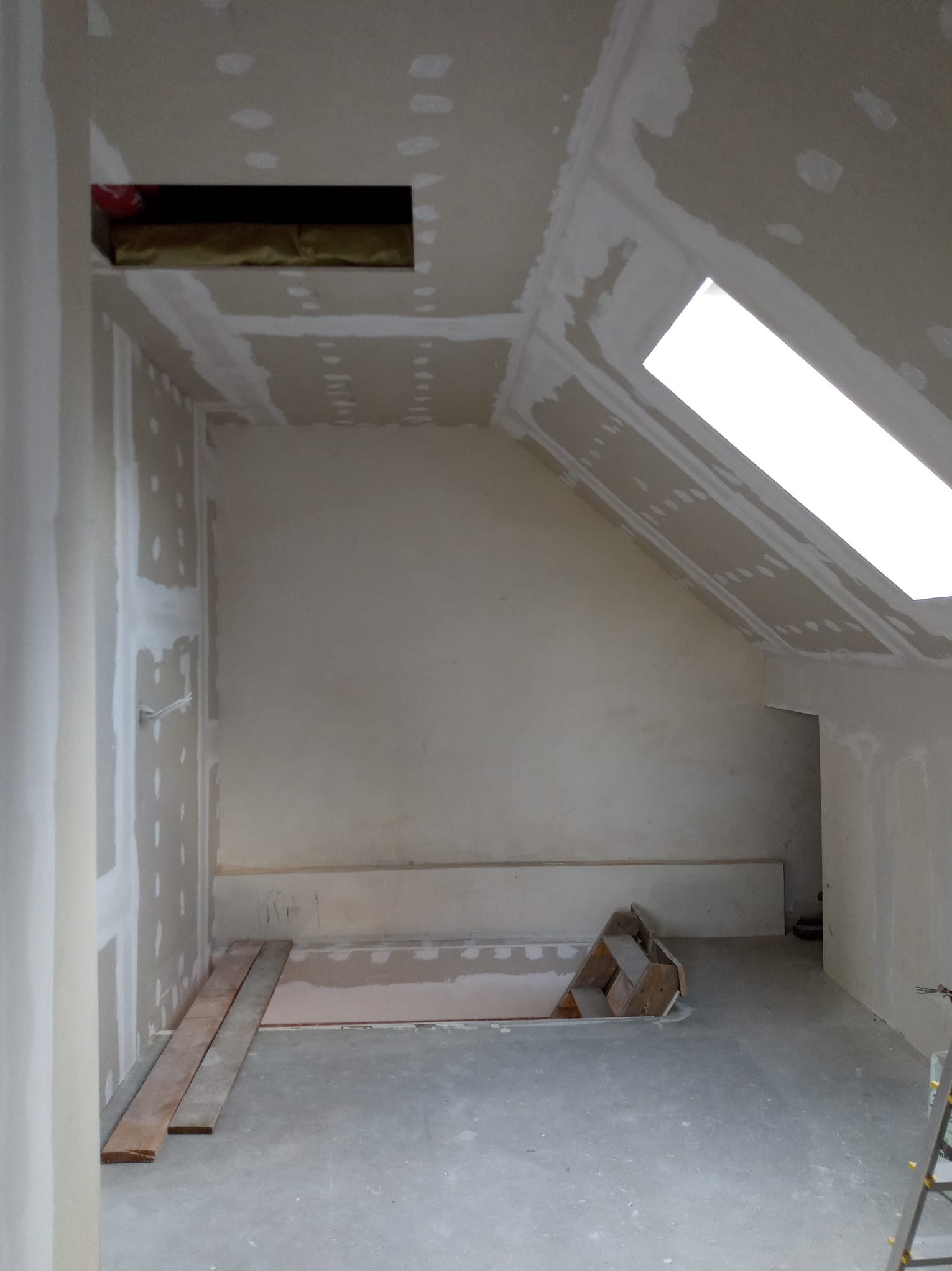 Poddasze w trakcie prac wykończeniowych: ściany i sufit pokryte płytami gipsowo-kartonowymi, widoczne szpachlowanie łączeń, okno dachowe, otwór w suficie z widoczną izolacją, fragment podłogi...