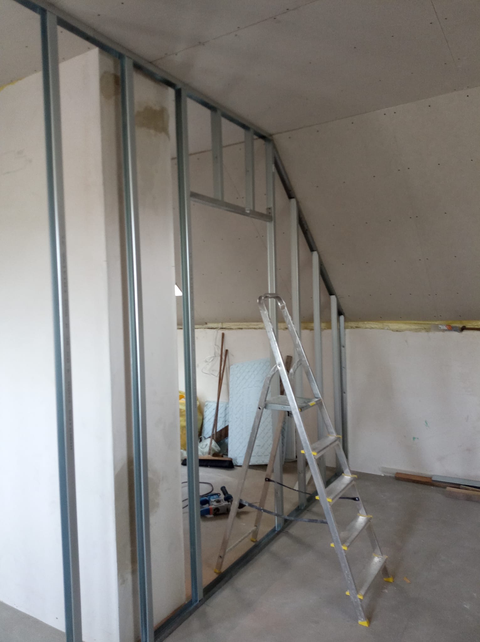 Stelaż z profili metalowych do montażu ścianki działowej z płyt gipsowo-kartonowych na poddaszu, widoczna drabina aluminiowa, narzędzia i materiały budowlane w tle.