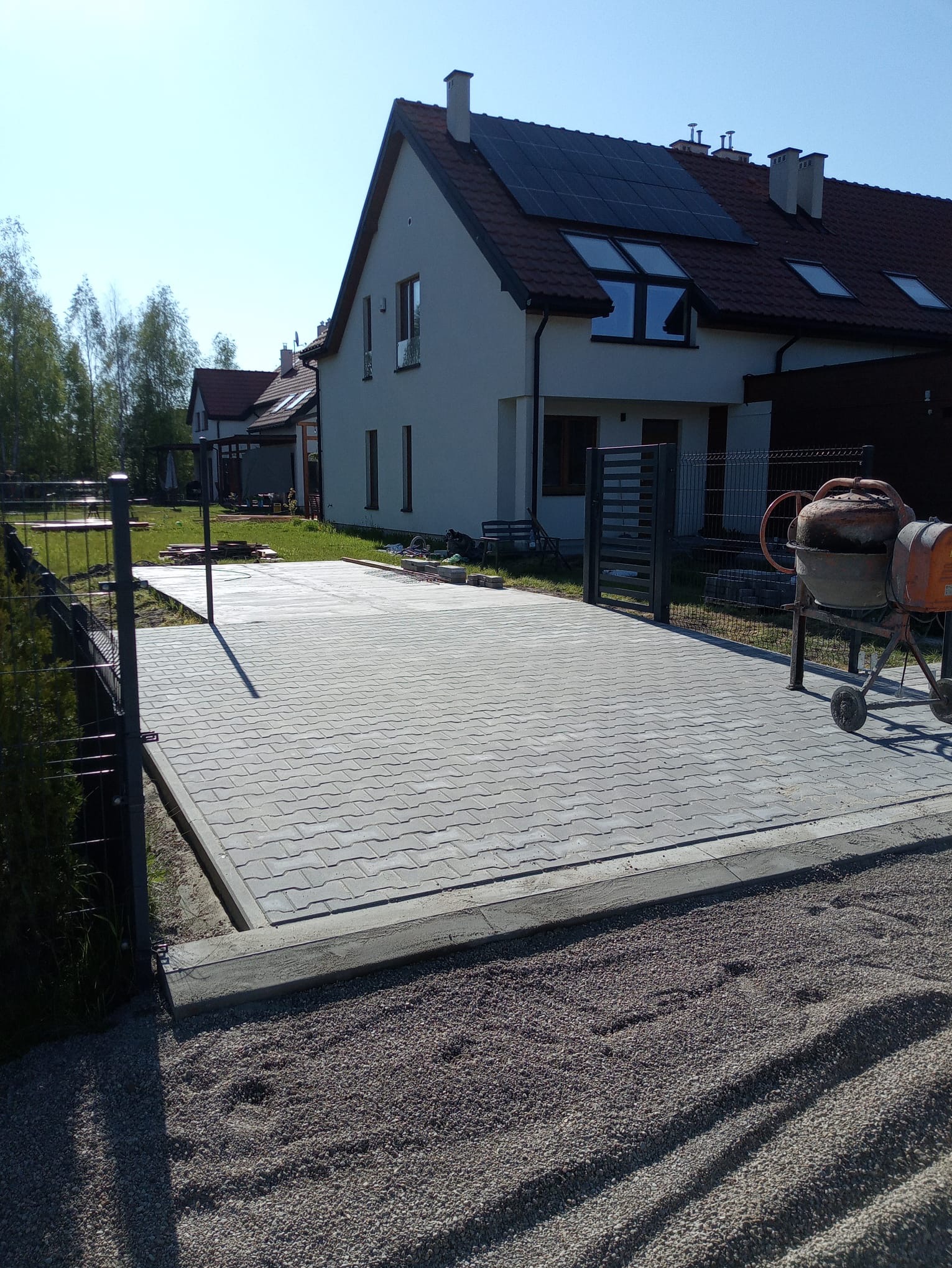 Ułożona kostka brukowa na podjeździe przed domem jednorodzinnym z panelami słonecznymi na dachu, widoczna betoniarka i przygotowane podłoże z kamyczków.