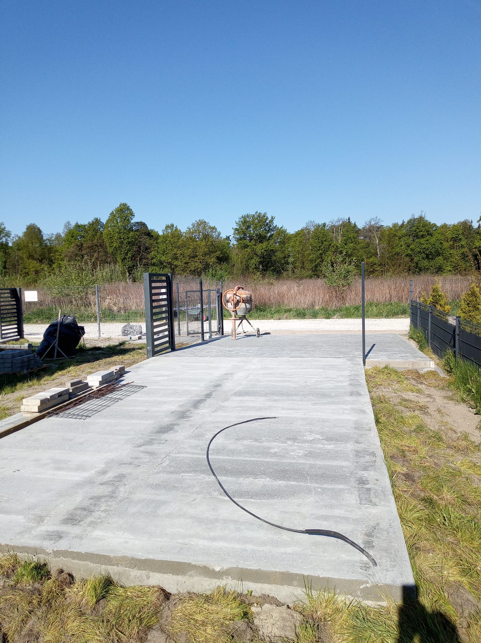 Betonowa podbudowa pod wjazd z przygotowanymi elementami ogrodzenia i bramy, widoczna betoniarka w tle, słoneczny dzień.