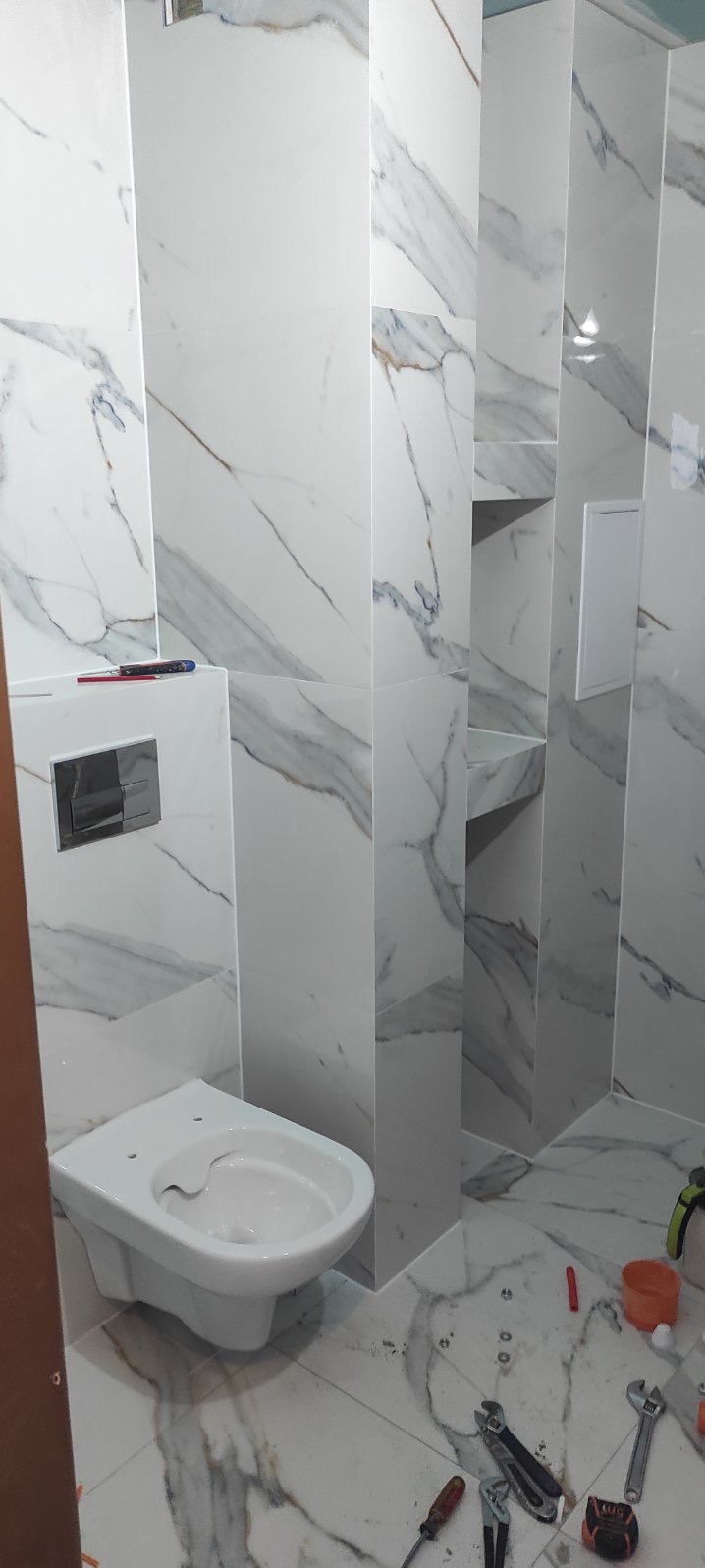 Montaż wiszącej toalety na tle marmurowych płytek, widoczne narzędzia glazurnicze na podłodze oraz wnęki w ścianie.