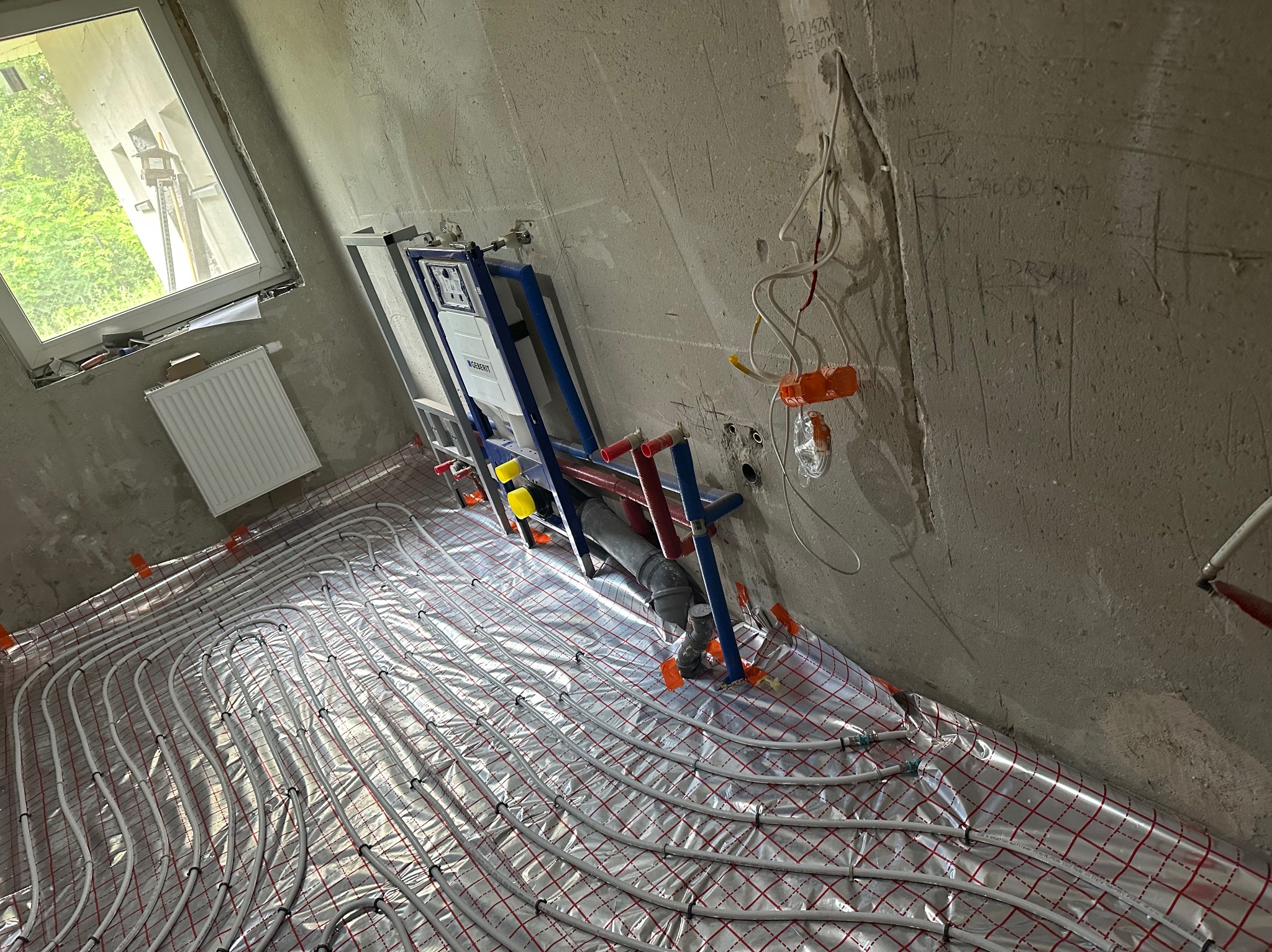 Instalacja ogrzewania podłogowego na folii z czerwoną siatką, widoczny stelaż podtynkowy WC Geberit na surowej ścianie z instalacją elektryczną i odręcznymi notatkami, okno w tle.