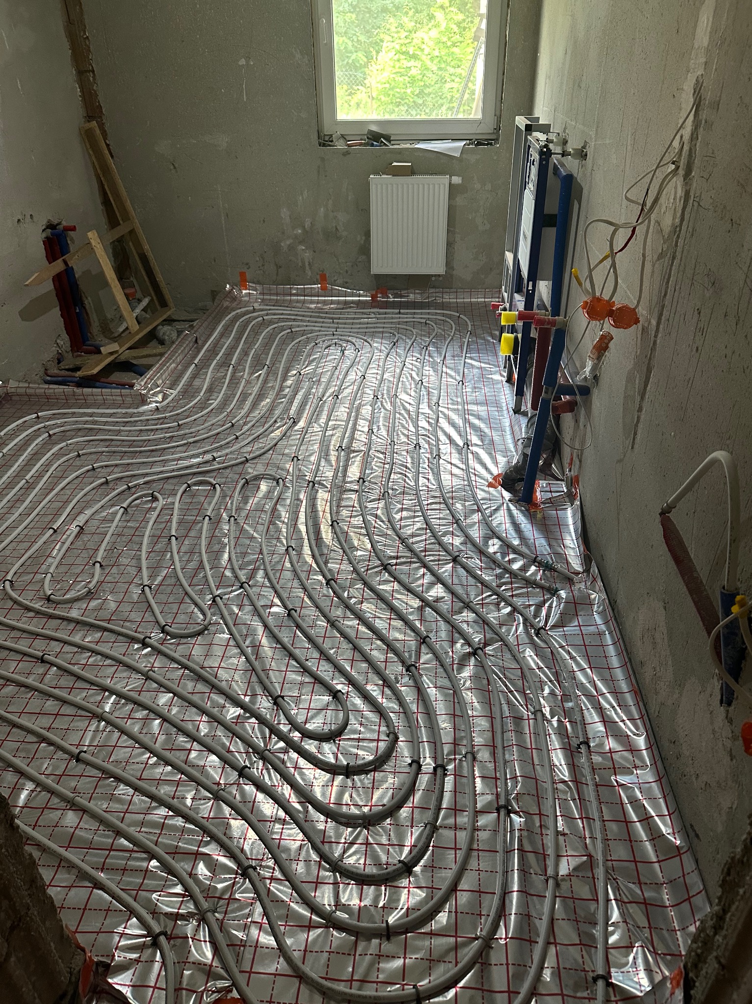 Instalacja ogrzewania podłogowego w surowym wnętrzu, rury grzewcze ułożone na folii z czerwoną kratką, widoczne elementy montażu podtynkowego WC i grzejnik pod oknem.