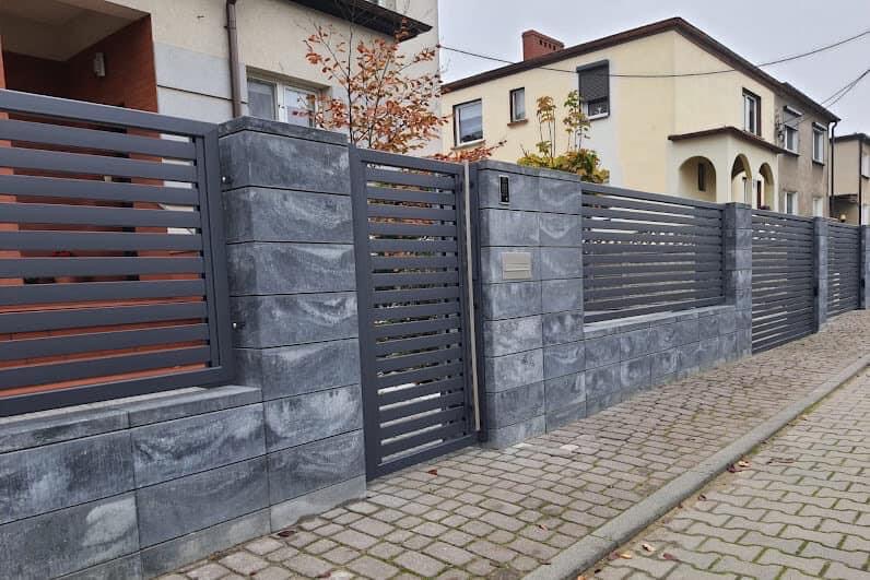 Nowoczesne ogrodzenie posesji z szarego kamienia łupanego i metalowych paneli w kolorze grafitowym, z furtką i skrzynką na listy, na tle dwupiętrowego domu.