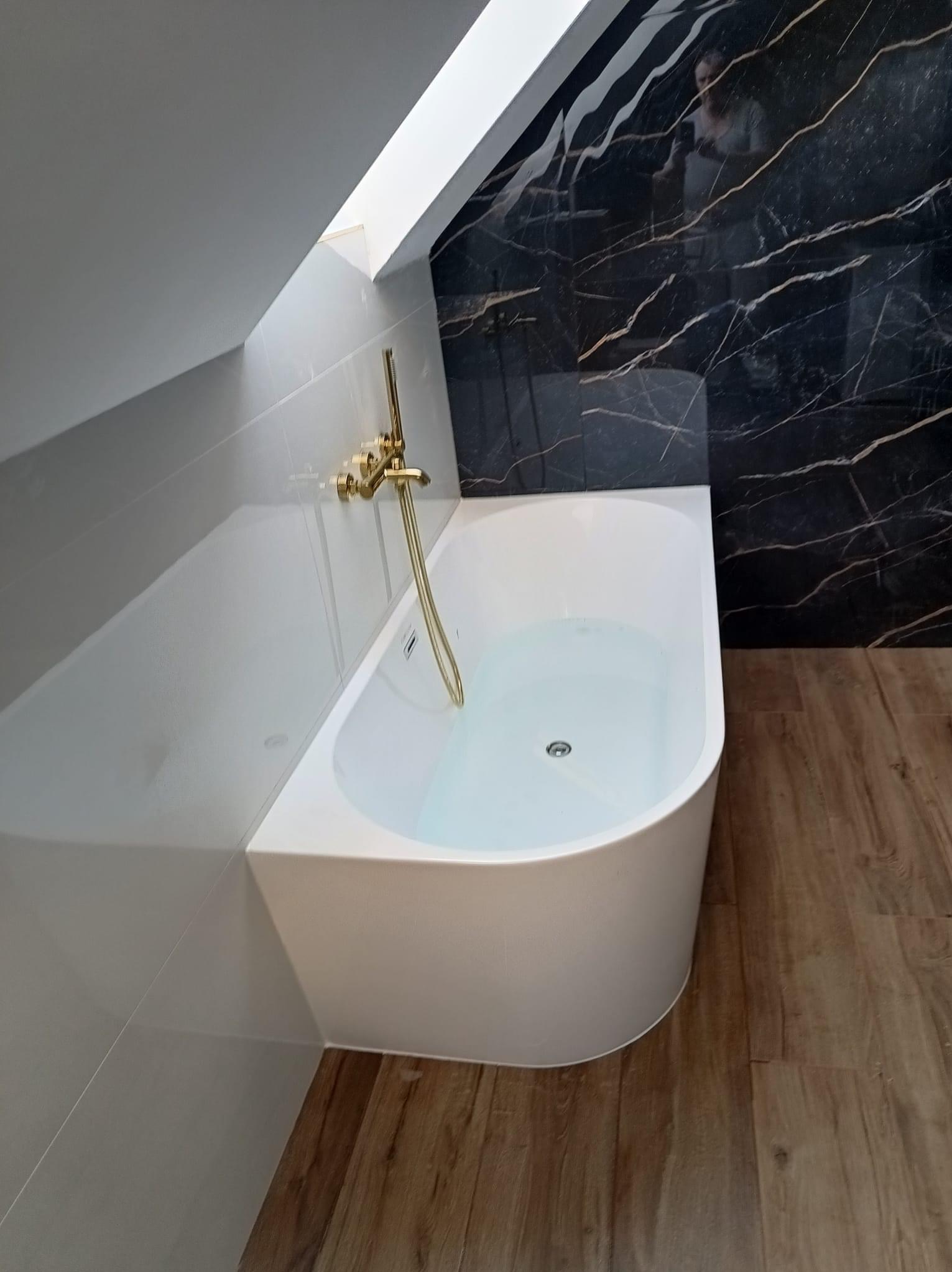 Biała wanna w łazience na poddaszu, złota bateria, ściana wyłożona czarnym marmurem z białymi żyłami, jasne panele podłogowe.