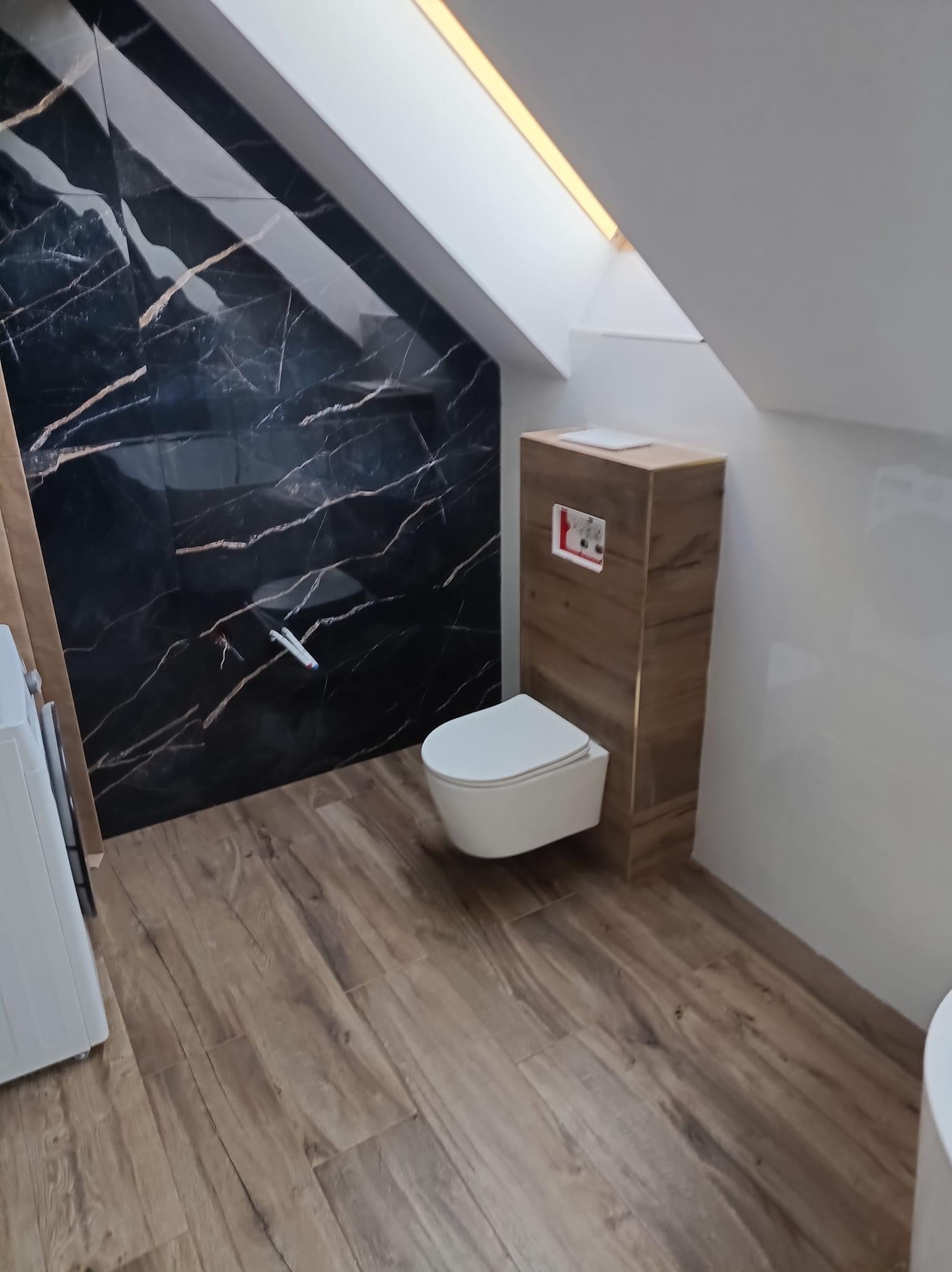 Nowoczesna łazienka na poddaszu z czarną marmurową ścianą, podwieszaną toaletą i drewnopodobną podłogą, oświetlona światłem z okna dachowego.