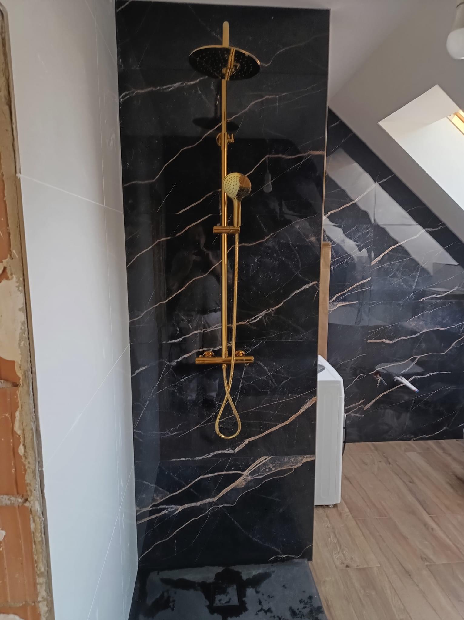 Nowoczesna łazienka w trakcie wykańczania: złota armatura prysznicowa na tle czarnych płytek imitujących marmur z wyraźnymi złotymi żyłami, widoczne białe płytki na ścianie obok i fragment surowej...
