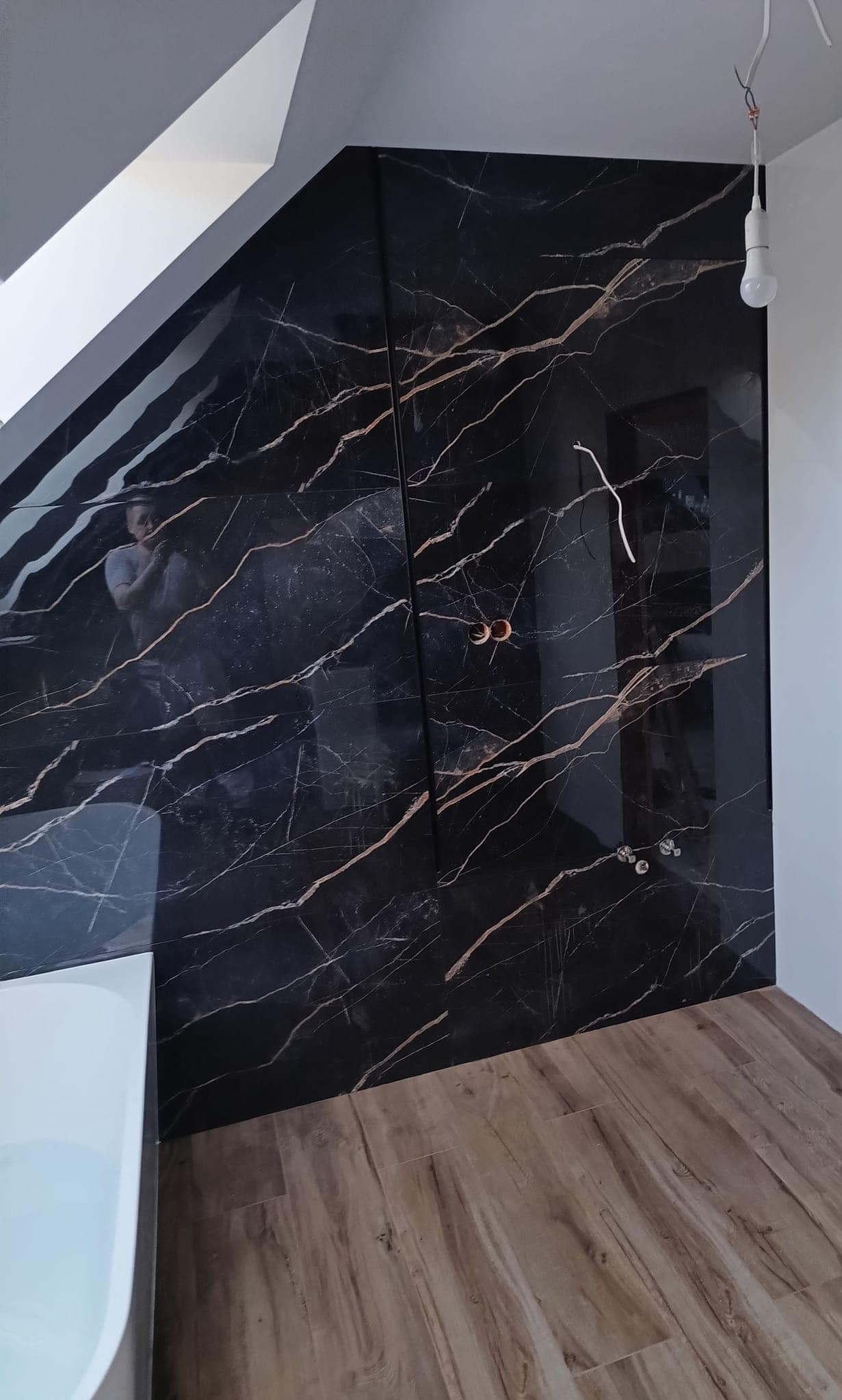 Ściana w łazience wyłożona czarnymi płytami z marmurowym wzorem, widoczne przygotowane punkty elektryczne, drewnopodobne panele podłogowe oraz fragment białej wanny.