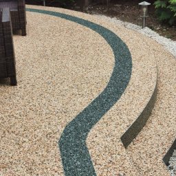 TPBUDOWA - Żwirowa ścieżka z ciemnym, żywicznym pasem biegnącym łagodnym łukiem, prowadząca do schodów wykończonych tym samym materiałem, otoczona zielenią i oświetlona niską lampą.