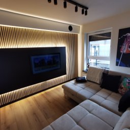 TPBUDOWA - Nowoczesny salon z drewnianą ścianą akcentującą z podświetleniem LED, zabudową TV w kolorze czarnym, jasną podłogą z paneli i narożnikiem w odcieniach beżu, uzupełniony o czarne reflektory sufitowe...