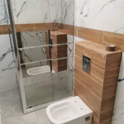 Nowoczesna łazienka w trakcie wykańczania: wisząca miska WC zamontowana obok obudowy stelaża z drewnopodobnych płytek, kabina prysznicowa ze szklanymi drzwiami, marmurowe płytki na ścianach, szare...