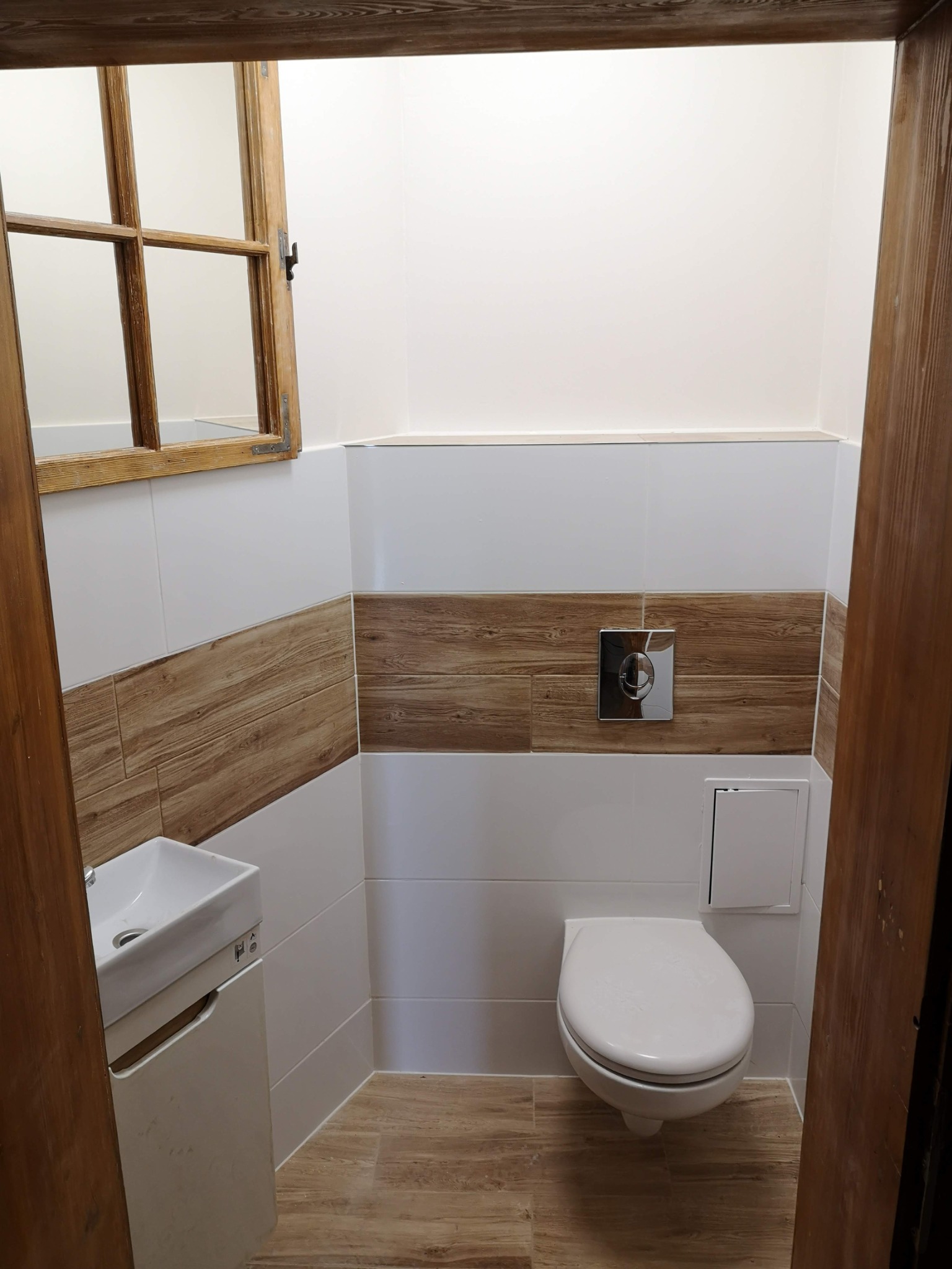 Mała toaleta z białymi i drewnopodobnymi płytkami, wiszącą muszlą klozetową, umywalką z szafką i drewnianym oknem w rustykalnym stylu.