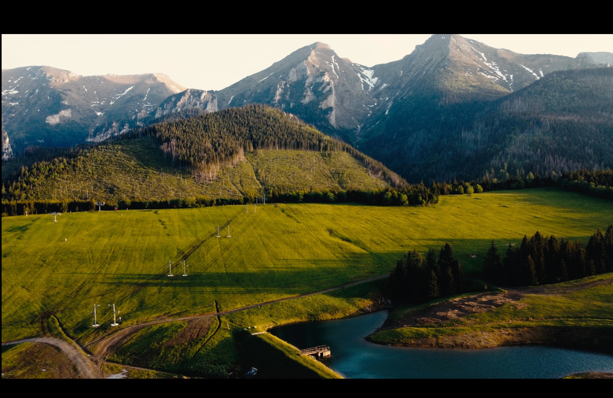 Panoramiczny widok z lotu ptaka na zieloną dolinę z jeziorem, otoczoną lasami i majestatycznymi szczytami górskimi ośnieżonymi na wierzchołkach, z widoczną infrastrukturą w postaci słupów i dróg.
