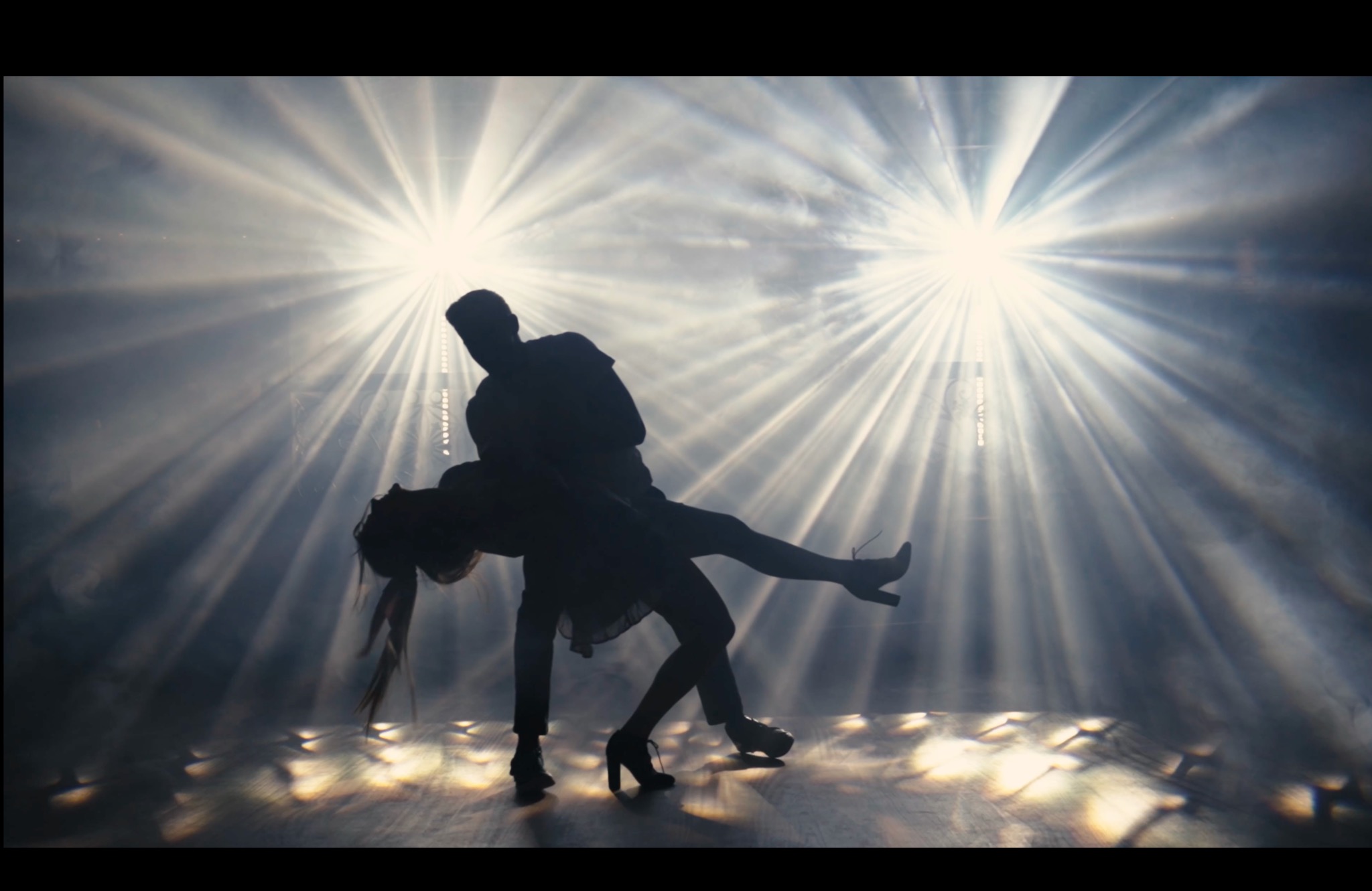 Sylwetki pary tanecznej w dynamicznej pozie, oświetlone intensywnymi reflektorami tworzącymi efektowne promienie światła i cienie na podłodze.