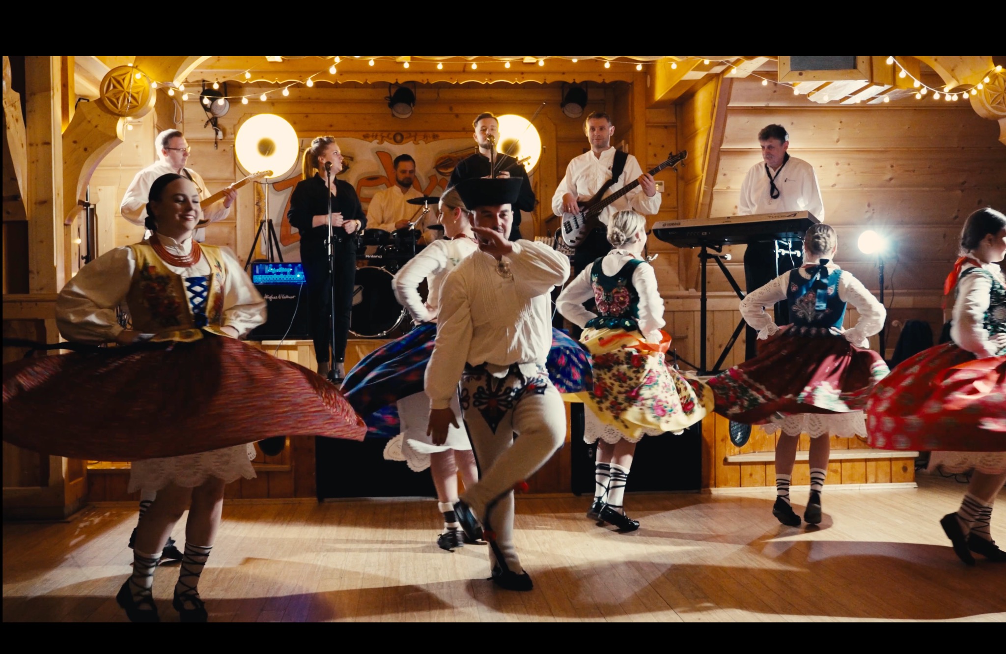 Zespół muzyczny i tancerze w strojach ludowych podczas występu na drewnianej scenie, oświetlonej lampami.