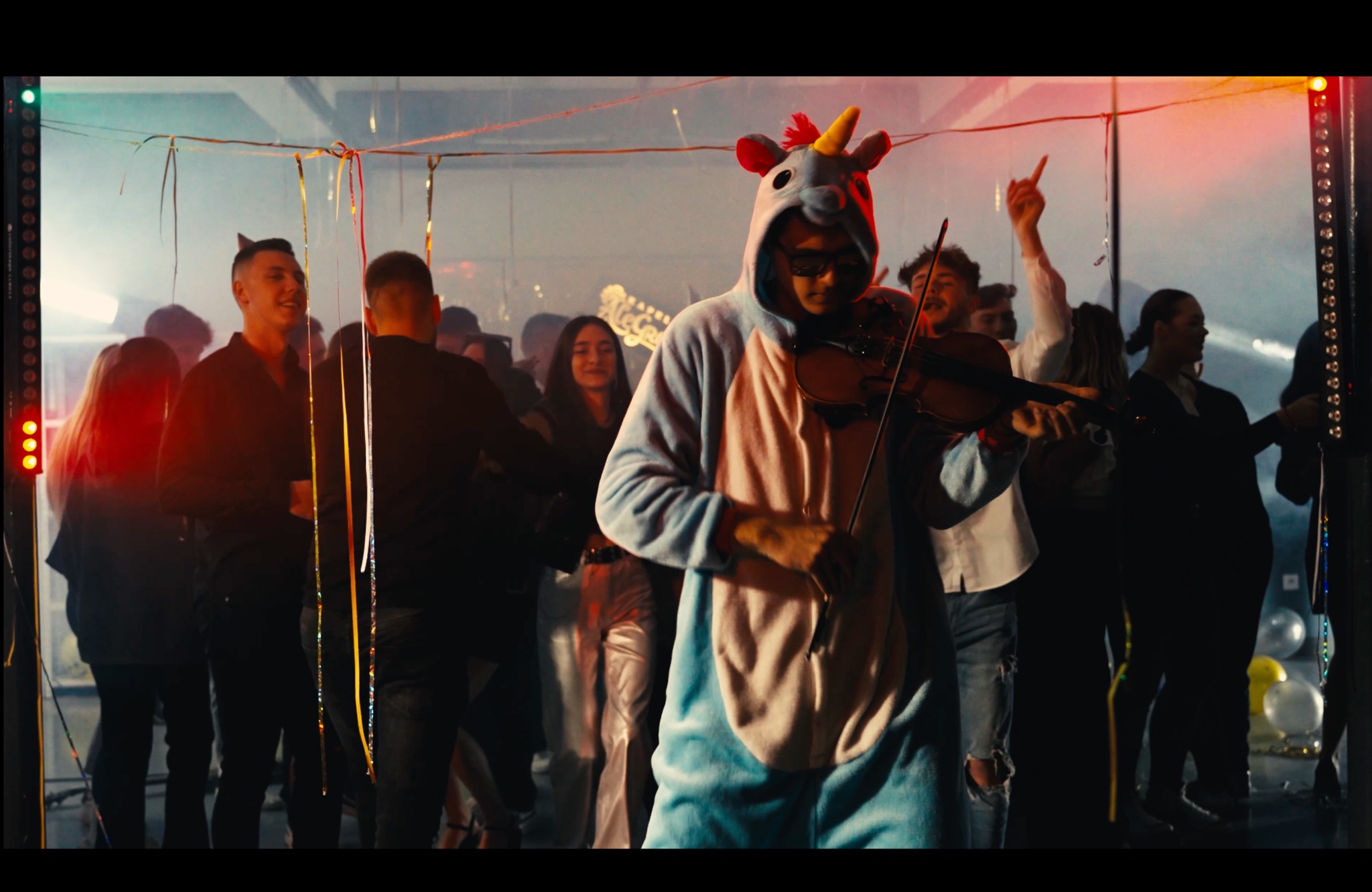 Osoba w kostiumie jednorożca grająca na skrzypcach w otoczeniu bawiących się ludzi, oświetlenie sceniczne i dekoracje imprezowe w tle.