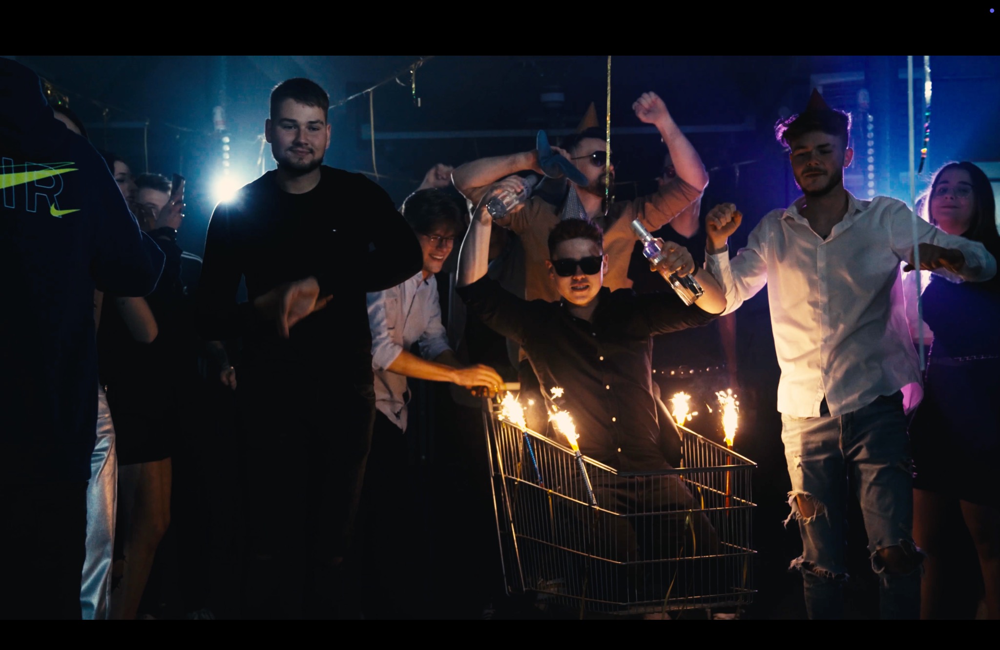 Mężczyzna w czarnej koszuli stoi w wózku sklepowym z płonącymi zimnymi ogniami, otoczony przez tańczących ludzi w słabym oświetleniu klubu.
