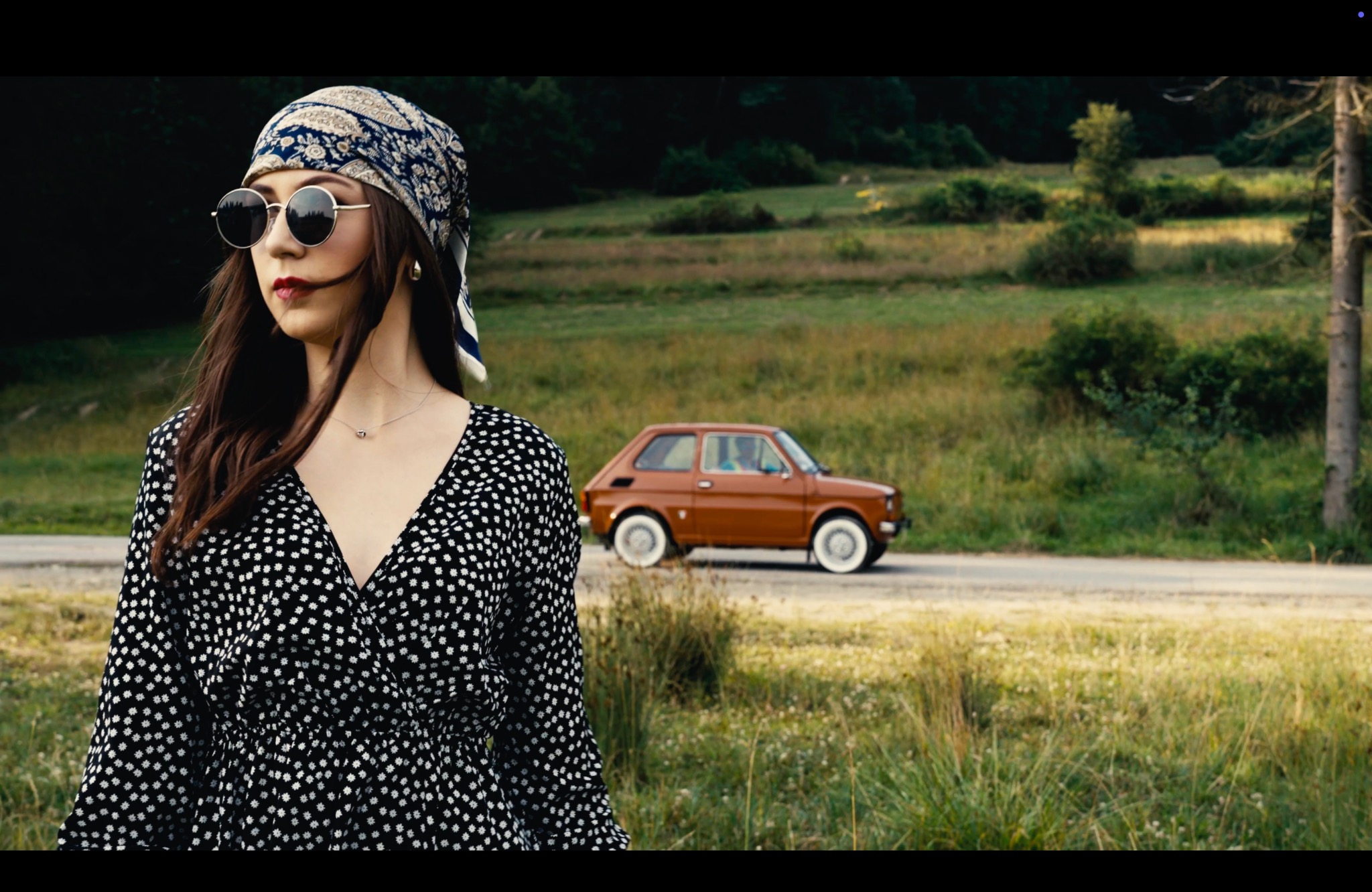 Kobieta w okularach przeciwsłonecznych i wzorzystej chuście, pozująca na tle krajobrazu z pomarańczowym samochodem.