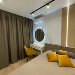 Nowoczesna sypialnia z beżowym łóżkiem, żółtymi poduszkami i krzesłem, oświetleniem szynowym i okrągłym lustrem z podświetleniem LED. Minimalistyczny design.