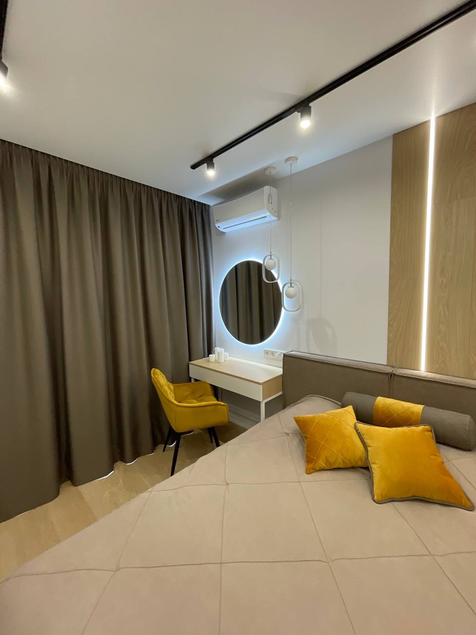 Nowoczesna sypialnia z beżowym łóżkiem, żółtymi poduszkami i krzesłem, oświetleniem szynowym i okrągłym lustrem z podświetleniem LED. Minimalistyczny design.