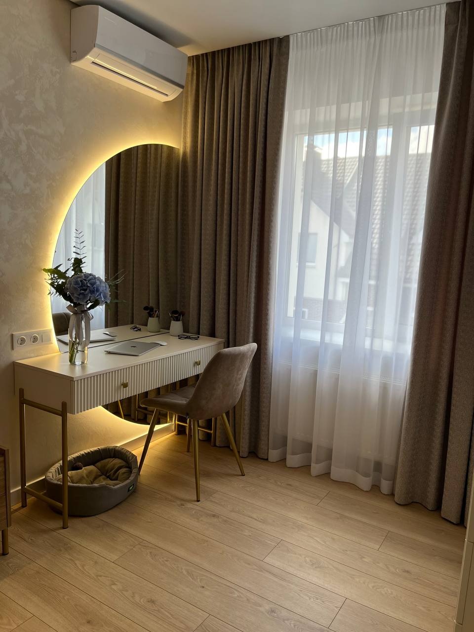 Elegancka toaletka z podświetlanym lustrem półksiężycowym, kwiatami, laptopem i beżowym krzesłem na tle okna z zasłonami i drewnianej podłogi.