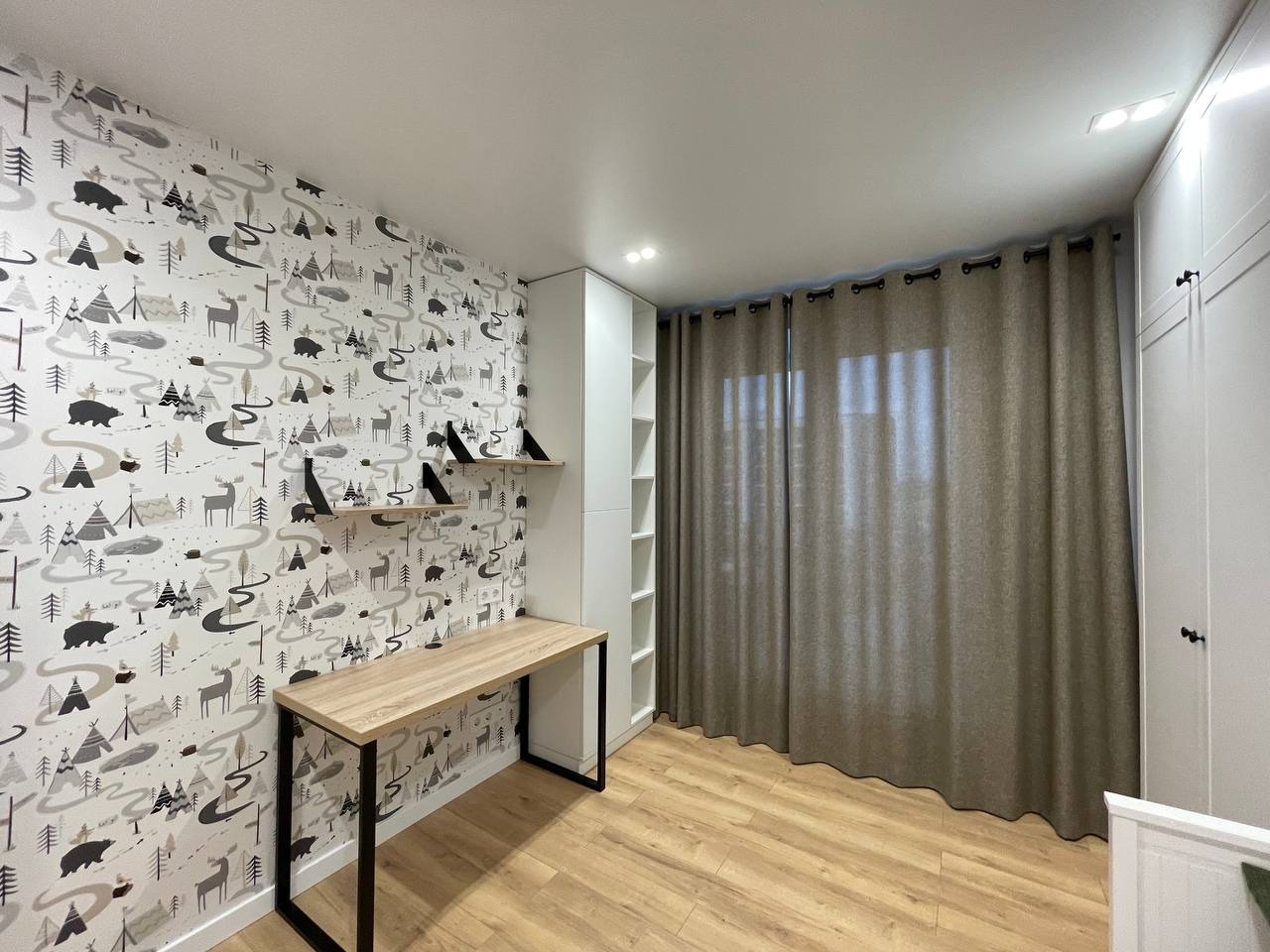 Pokój dziecięcy z tapetą w leśny motyw, biurkiem na metalowych nogach, drewnianą podłogą i zasłonami w kolorze beżowym. Widoczne półki i szafa w zabudowie.