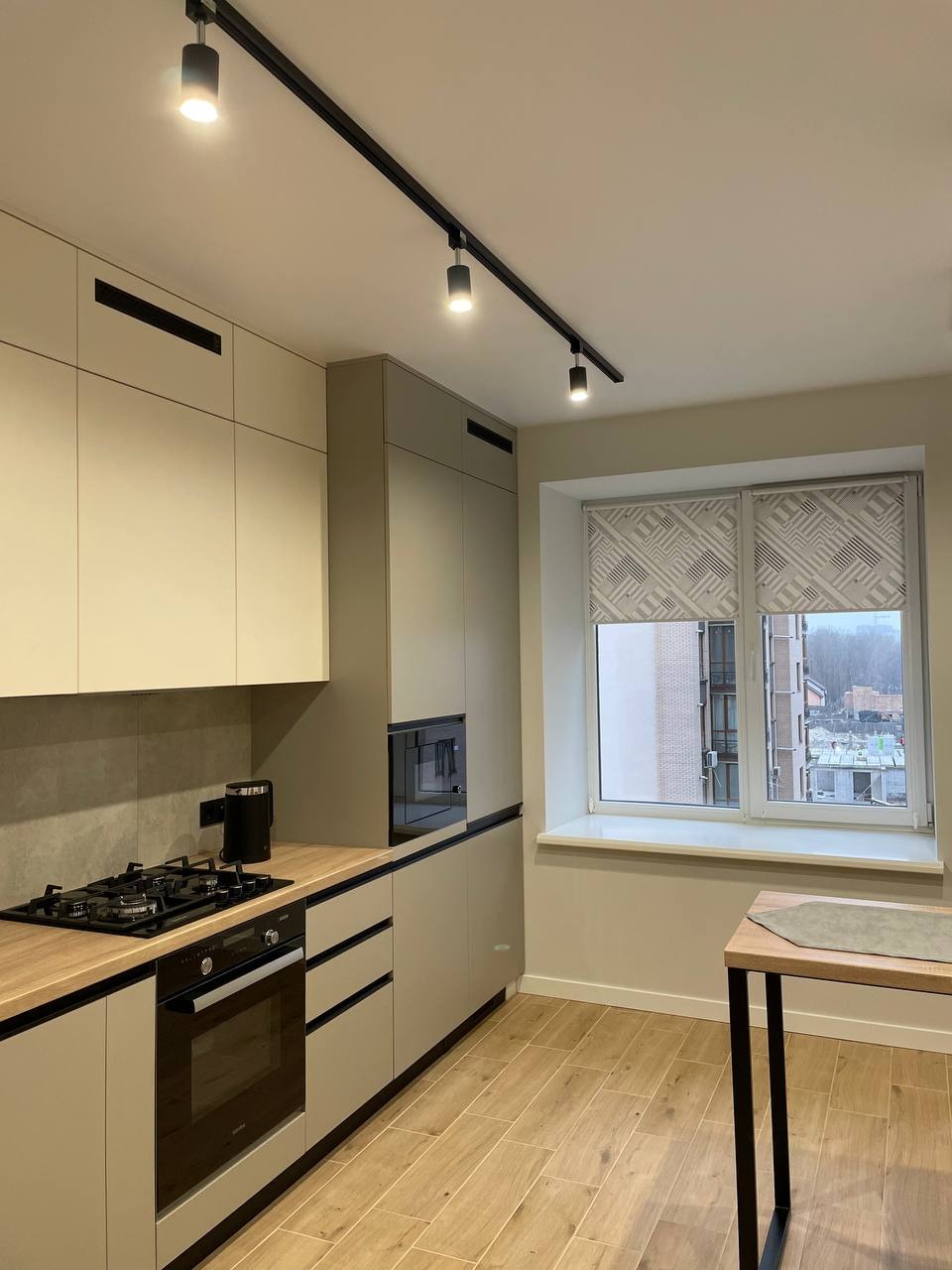 Jasna kuchnia z drewnianym blatem, czarną płytą grzewczą i oświetleniem szynowym. Okno z roletą w tle. Nowoczesne wykończenie wnętrza.
