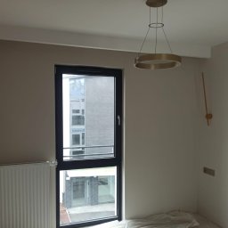 ProBud Group - Pomalowane na beżowo wnętrze z grzejnikiem, oknem z czarną ramą i nowoczesną lampą wiszącą w kształcie okręgu, podłoga zabezpieczona folią malarską.