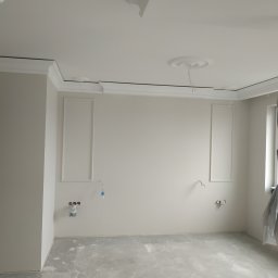 ProBud Group - Pokój w trakcie remontu: ściany z dekoracyjnymi panelami i listwami, zabezpieczone instalacje elektryczne, podłoga z wylewką, okno osłonięte folią malarską.