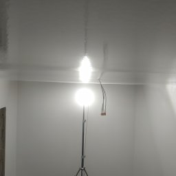ProBud Group - Świeżo pomalowany, błyszczący sufit w pokoju, oświetlony lampą budowlaną na statywie, widoczne kable lampy, przygotowane do dalszych prac wykończeniowych, stan surowy w narożnikach.