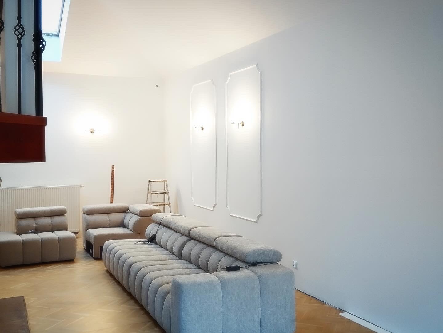 Jasne wnętrze po remoncie z gładkimi, białymi ścianami, dekoracyjnymi listwami ściennymi, minimalistycznym oświetleniem i nowoczesnym, modułowym narożnikiem na parkiecie.