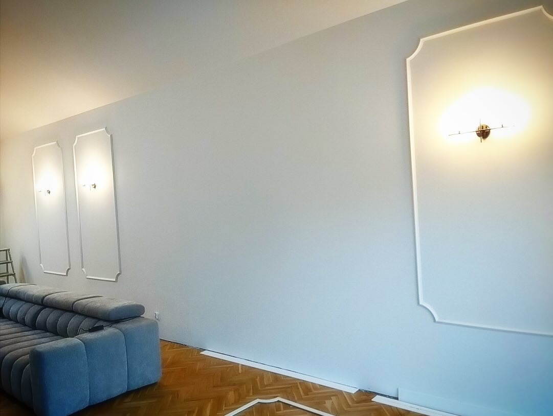 Jasnoszary salon po remoncie z dekoracyjnymi białymi listwami ściennymi, parkietem i szarą sofą modułową.