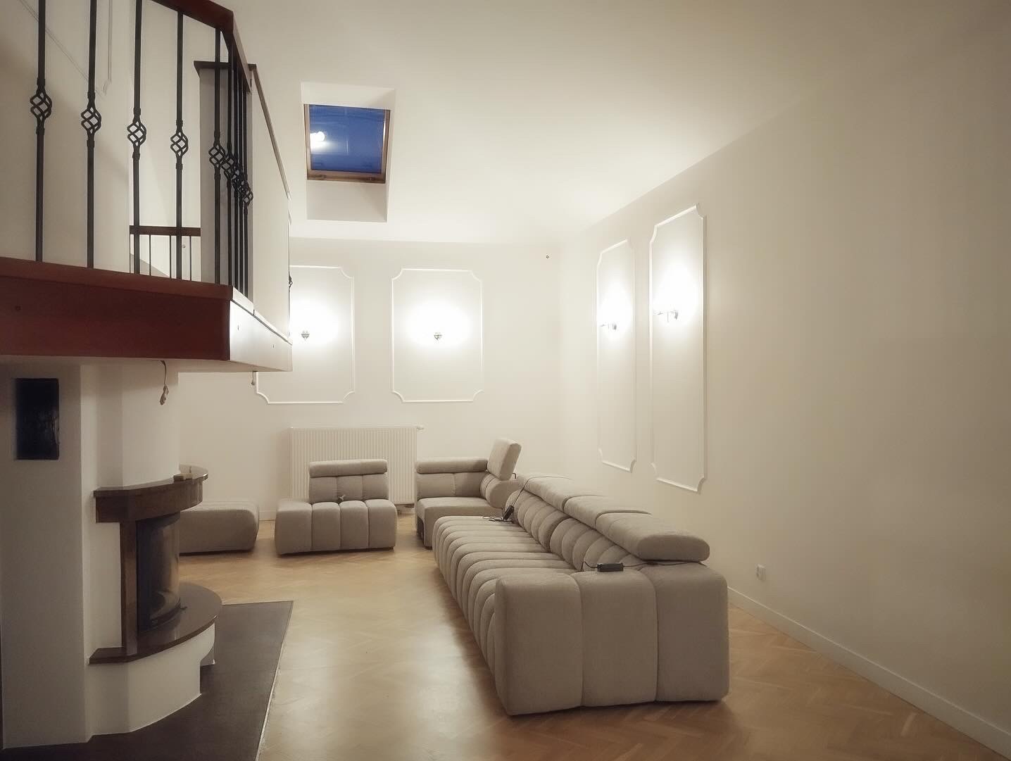 Jasne wnętrze salonu po remoncie: kremowe ściany z dekoracyjnymi listwami, parkiet w jodełkę, nowoczesny kominek pod schodami z kutą balustradą oraz minimalistyczne meble wypoczynkowe.