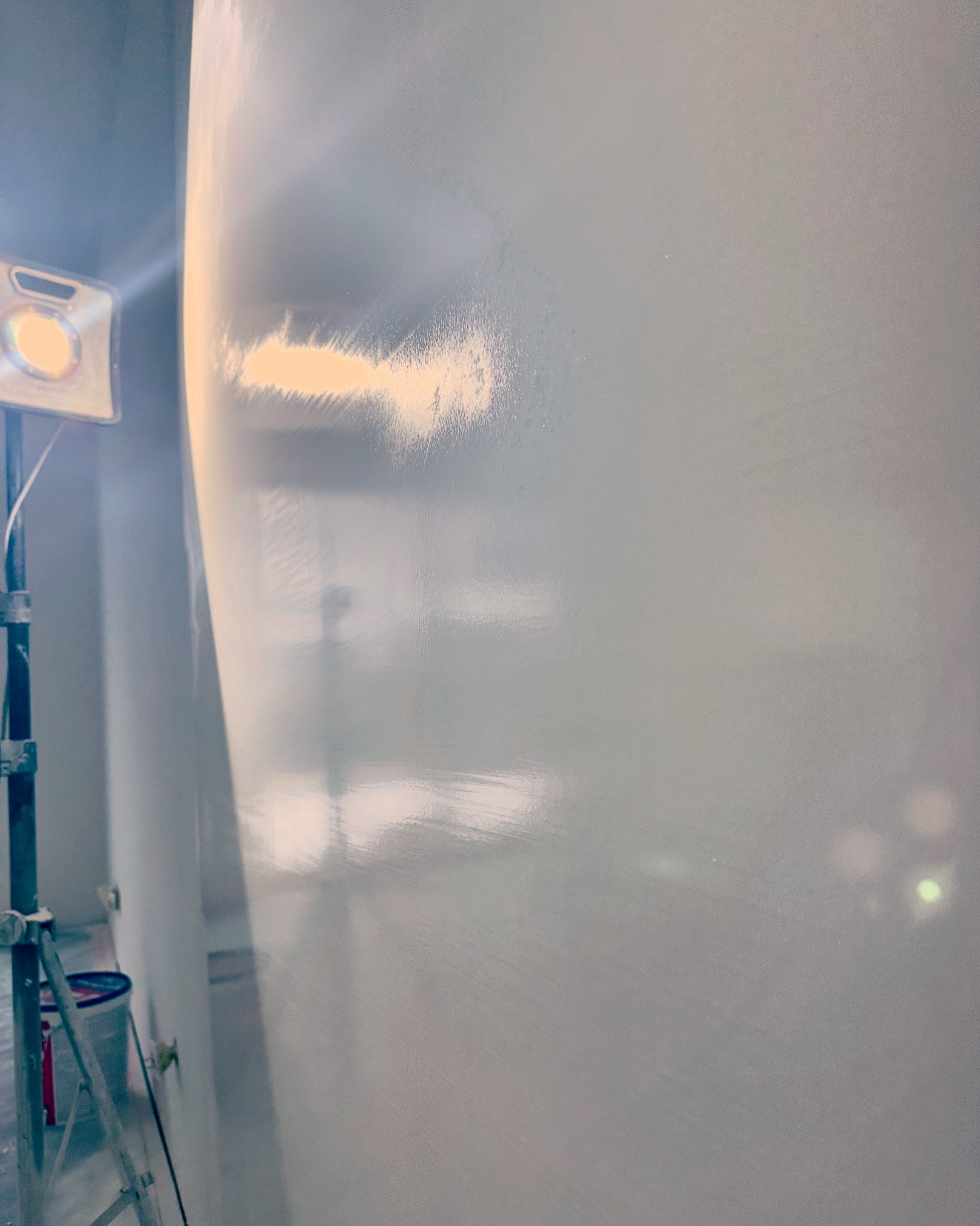 Błyszcząca, świeżo pomalowana ściana w odcieniach szarości, oświetlona lampą budowlaną na statywie, widoczny fragment drabiny malarskiej i wiaderka z farbą.