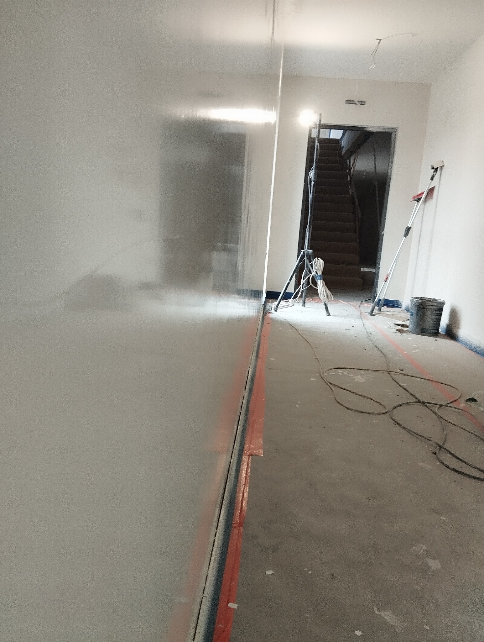 Świeżo pomalowana, błyszcząca ściana w trakcie remontu, z widocznymi śladami taśmy malarskiej na podłodze, drabina i narzędzia malarskie w tle.