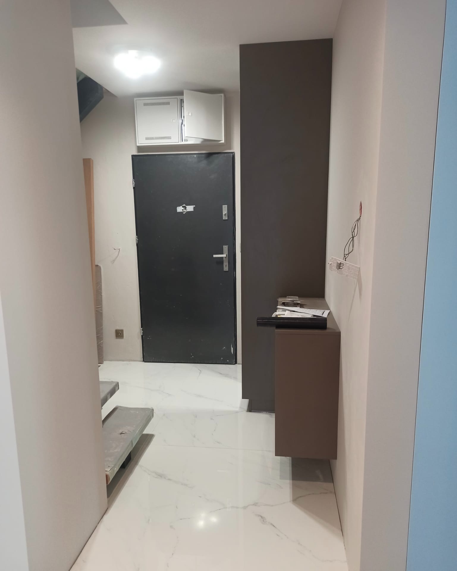 Korytarz po remoncie z białymi ścianami, marmurową podłogą i ciemnymi akcentami: drzwi wejściowe, panel na ścianie i szafka, widoczne zabezpieczone schody