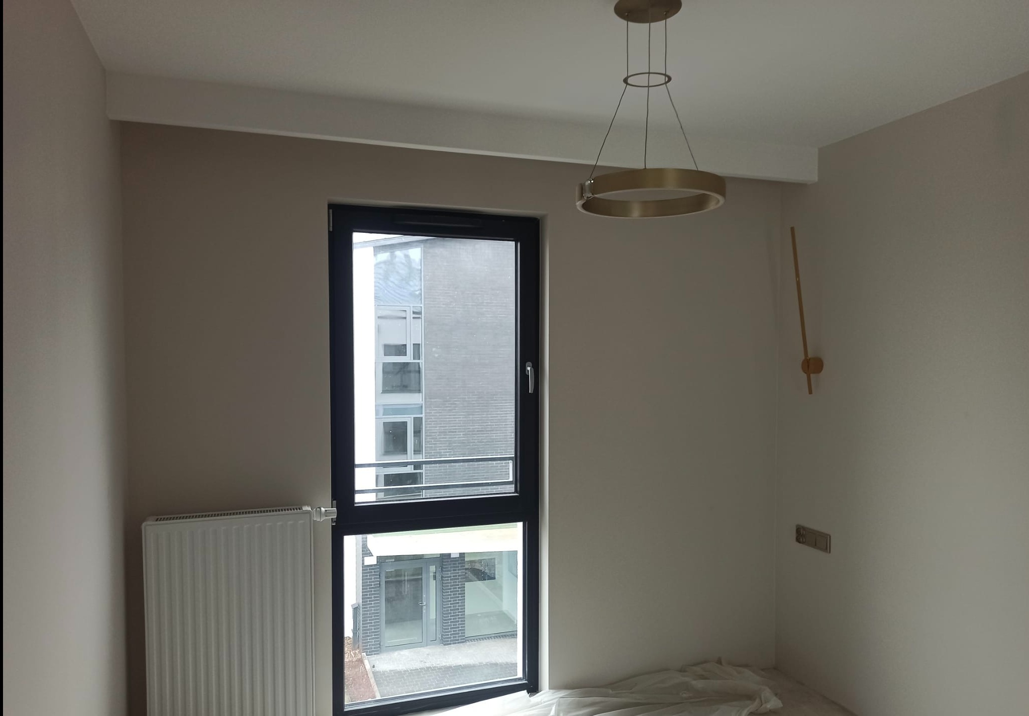 Pomalowane na beżowo wnętrze z grzejnikiem, oknem z czarną ramą i nowoczesną lampą wiszącą w kształcie okręgu, podłoga zabezpieczona folią malarską.
