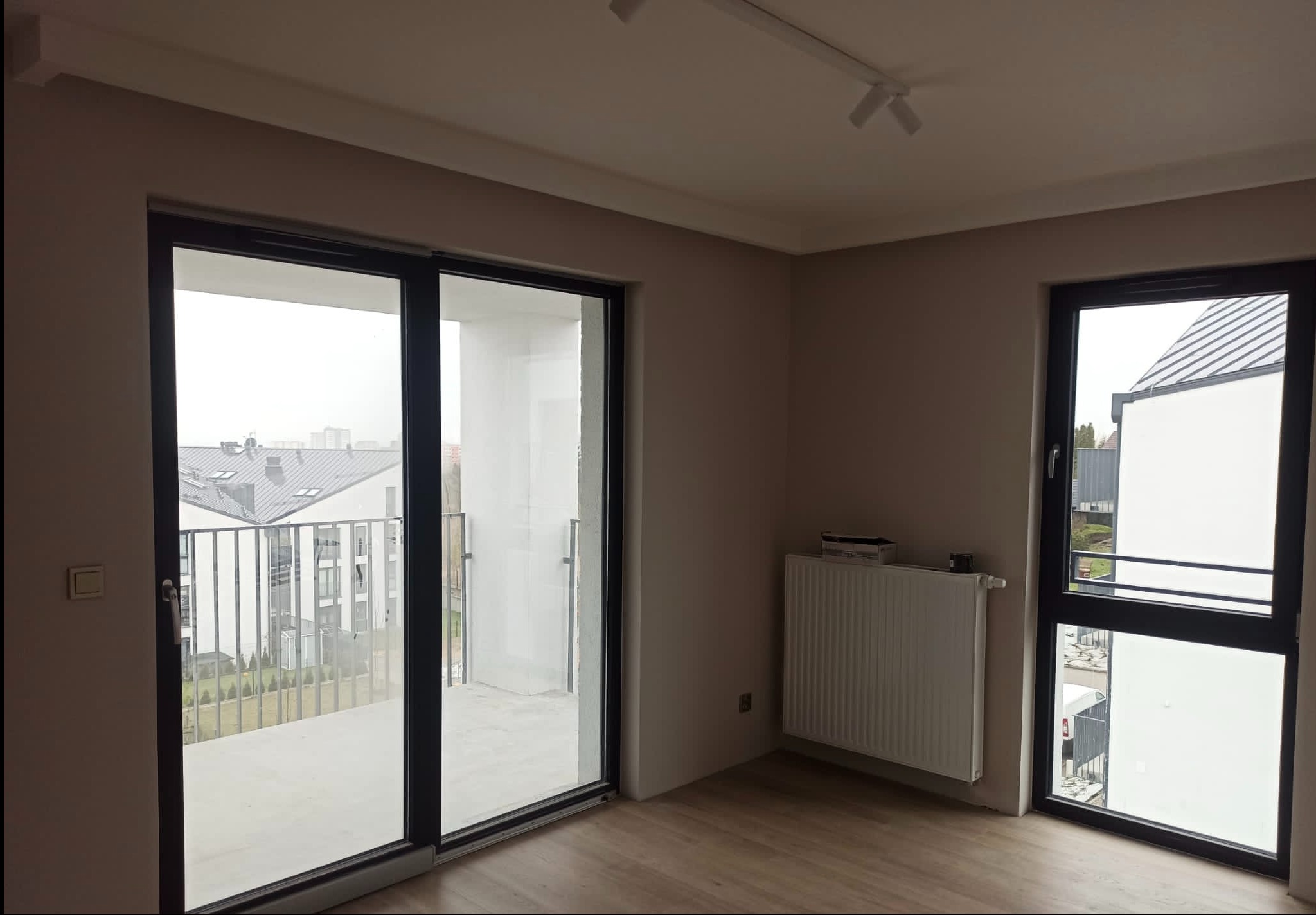 Wnętrze nowego mieszkania z beżowymi ścianami, dużym oknem balkonowym z widokiem na balkon i osiedle, grzejnikiem pod oknem oraz drewnopodobną podłogą.