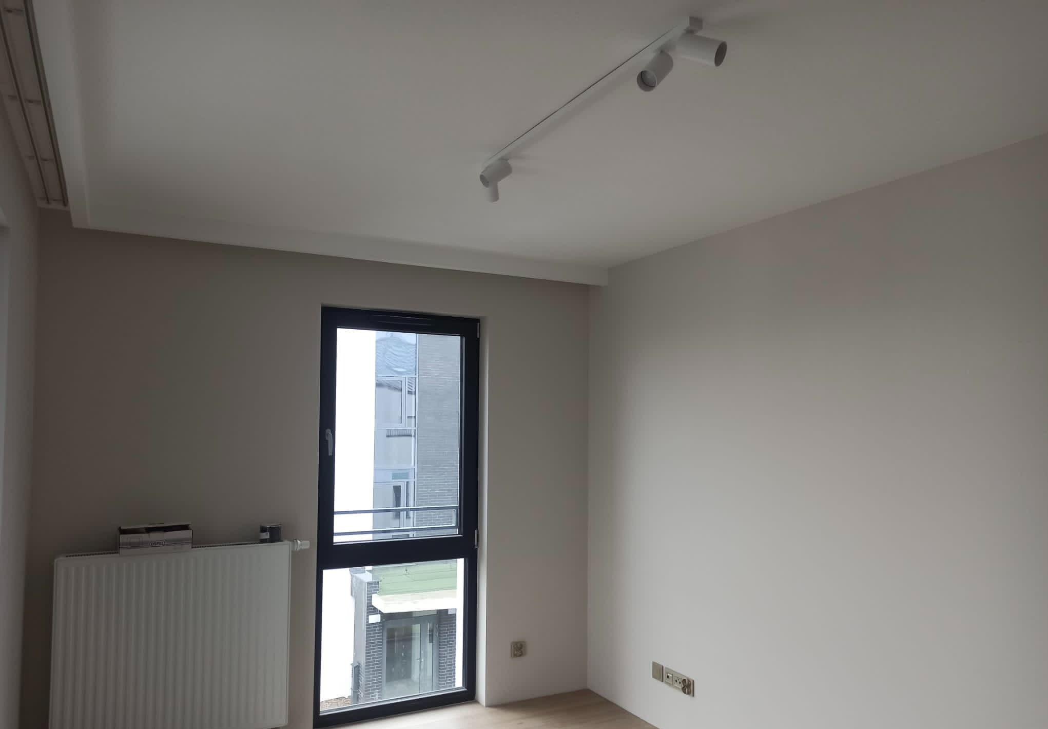 Wnętrze świeżo wykończonego pokoju z pomalowanymi ścianami na jasny odcień szarości, widoczne okno z czarną ramą, biały grzejnik pod oknem oraz nowoczesne oświetlenie sufitowe na szynie.