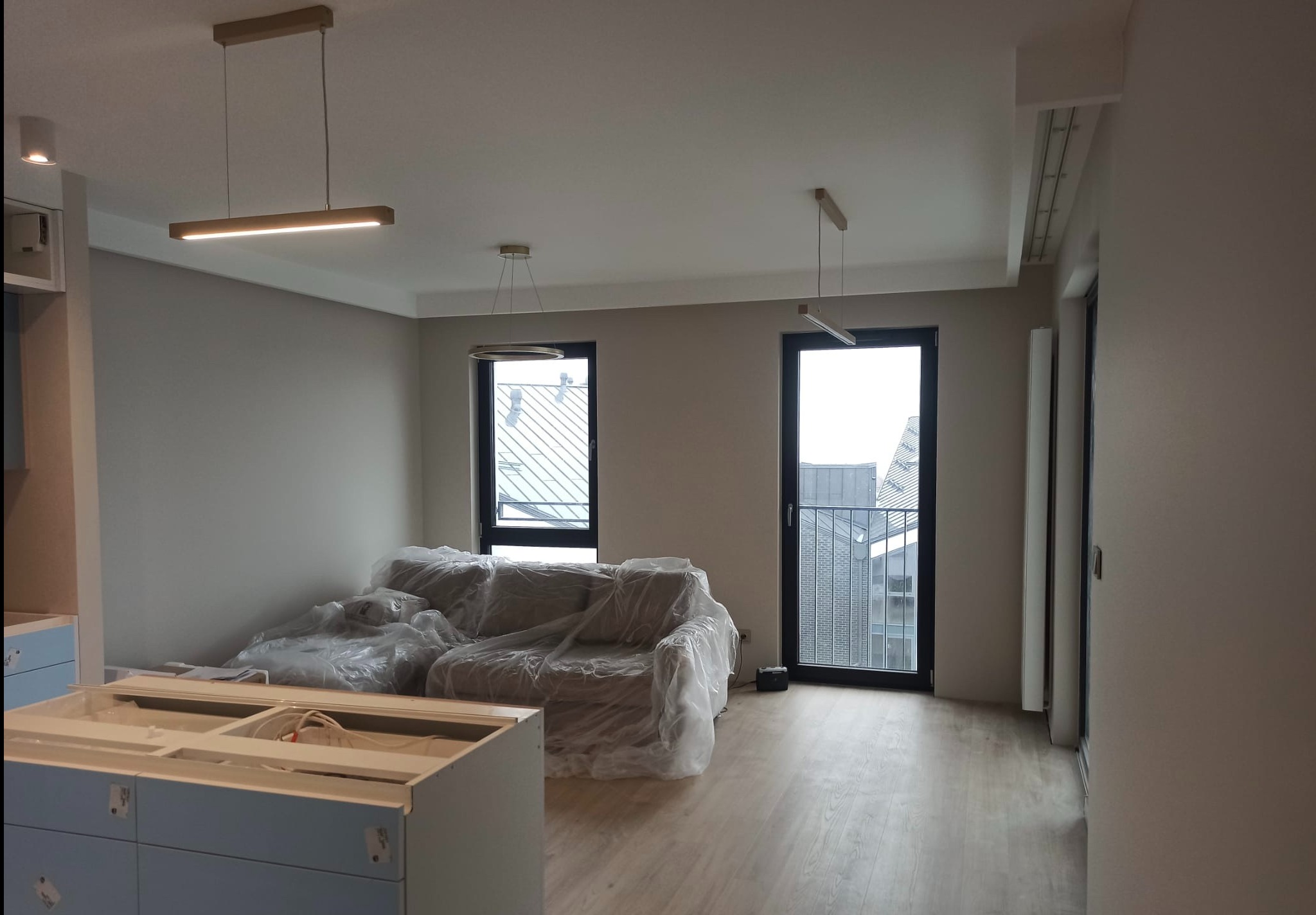 Pokój w trakcie prac wykończeniowych: ściany po malowaniu na neutralny kolor, sofa zabezpieczona folią, widoczne nowoczesne lampy sufitowe, podłoga wyłożona panelami, w tle okna z widokiem na miasto.