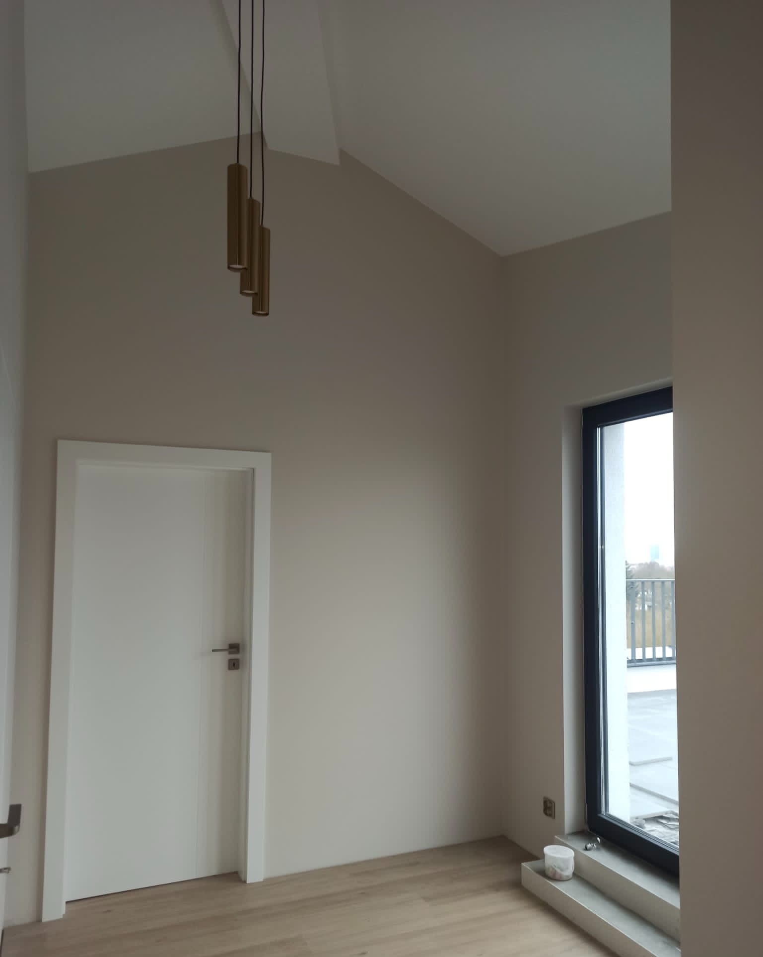 Wnętrze po odświeżeniu w Szczecinie: kremowe ściany, białe drzwi i okno z czarną ramą, nowoczesna lampa wisząca z trzema złotymi tubami.