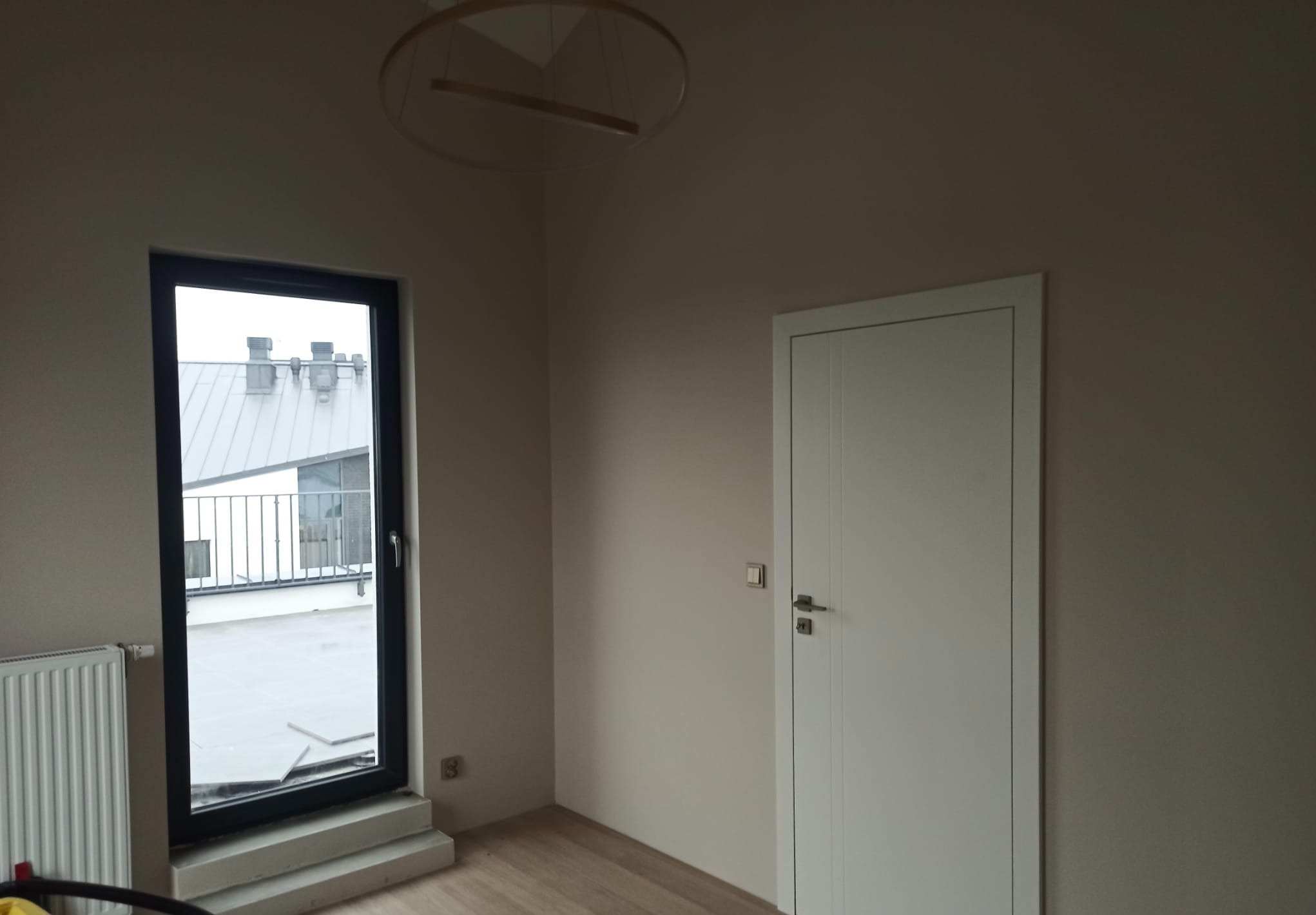 Pomalowane na beżowo wnętrze z białymi drzwiami i grzejnikiem, nowoczesna lampa sufitowa, okno z widokiem na dach.
