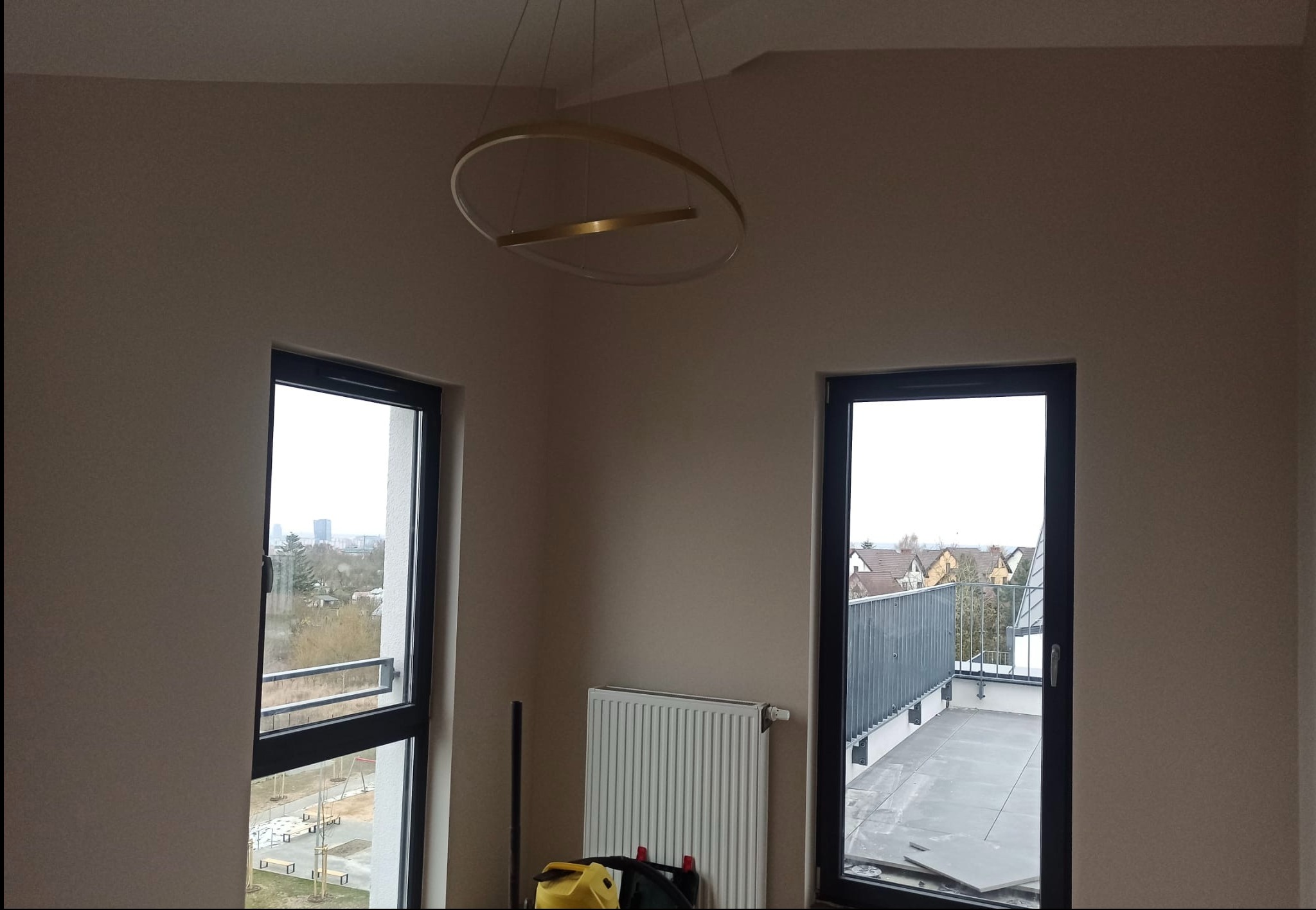 Wnętrze pokoju po remoncie z dwoma oknami i wyjściem na balkon, ściany w odcieniach beżu, nowoczesna lampa sufitowa, widoczny grzejnik i sprzęt do sprzątania.
