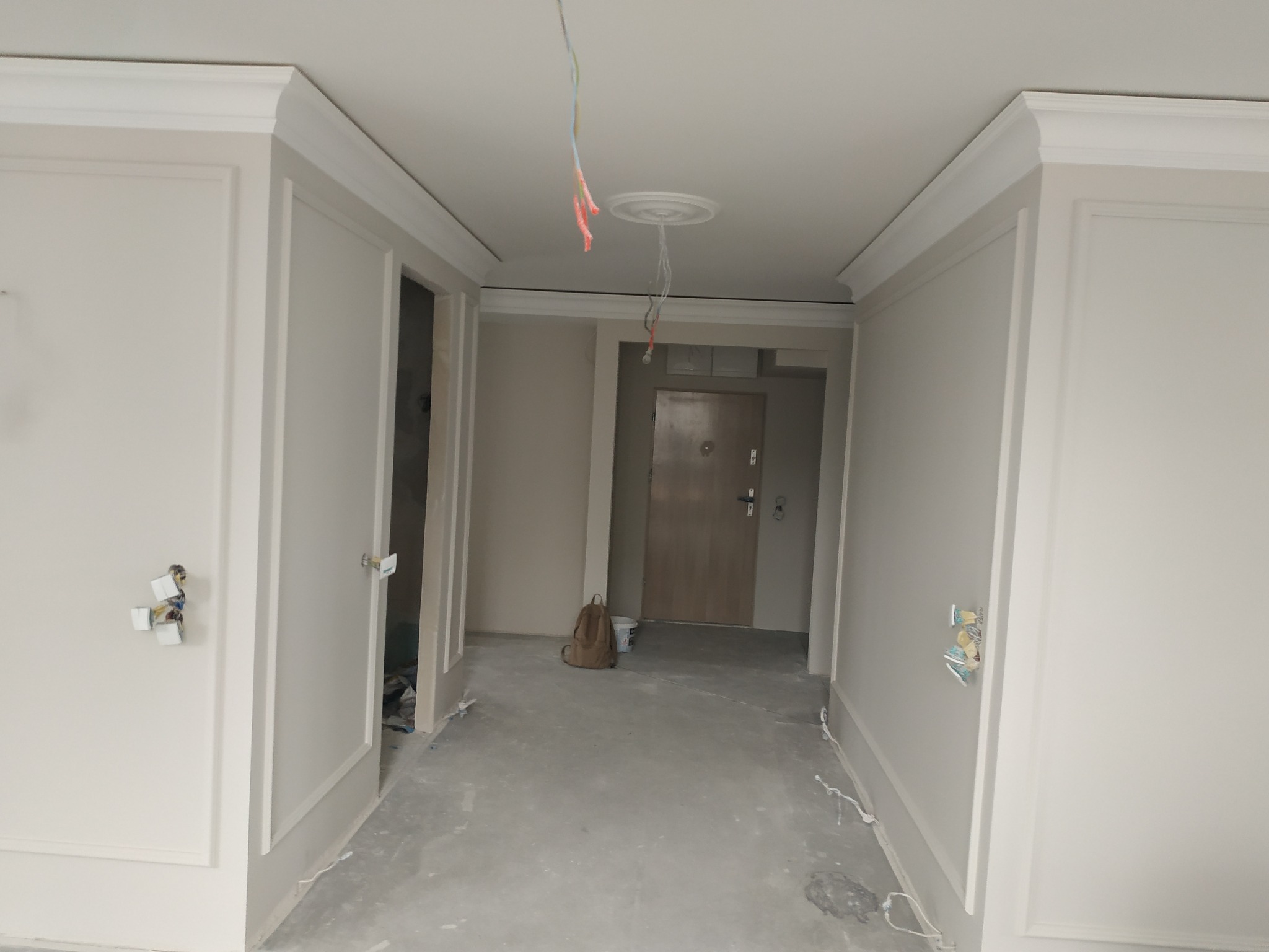 Korytarz w trakcie remontu: ściany z dekoracyjnymi panelami pomalowane na jasny odcień, surowy beton na podłodze, widoczne wystające kable i puszki elektryczne, w głębi drzwi wejściowe i plecak...