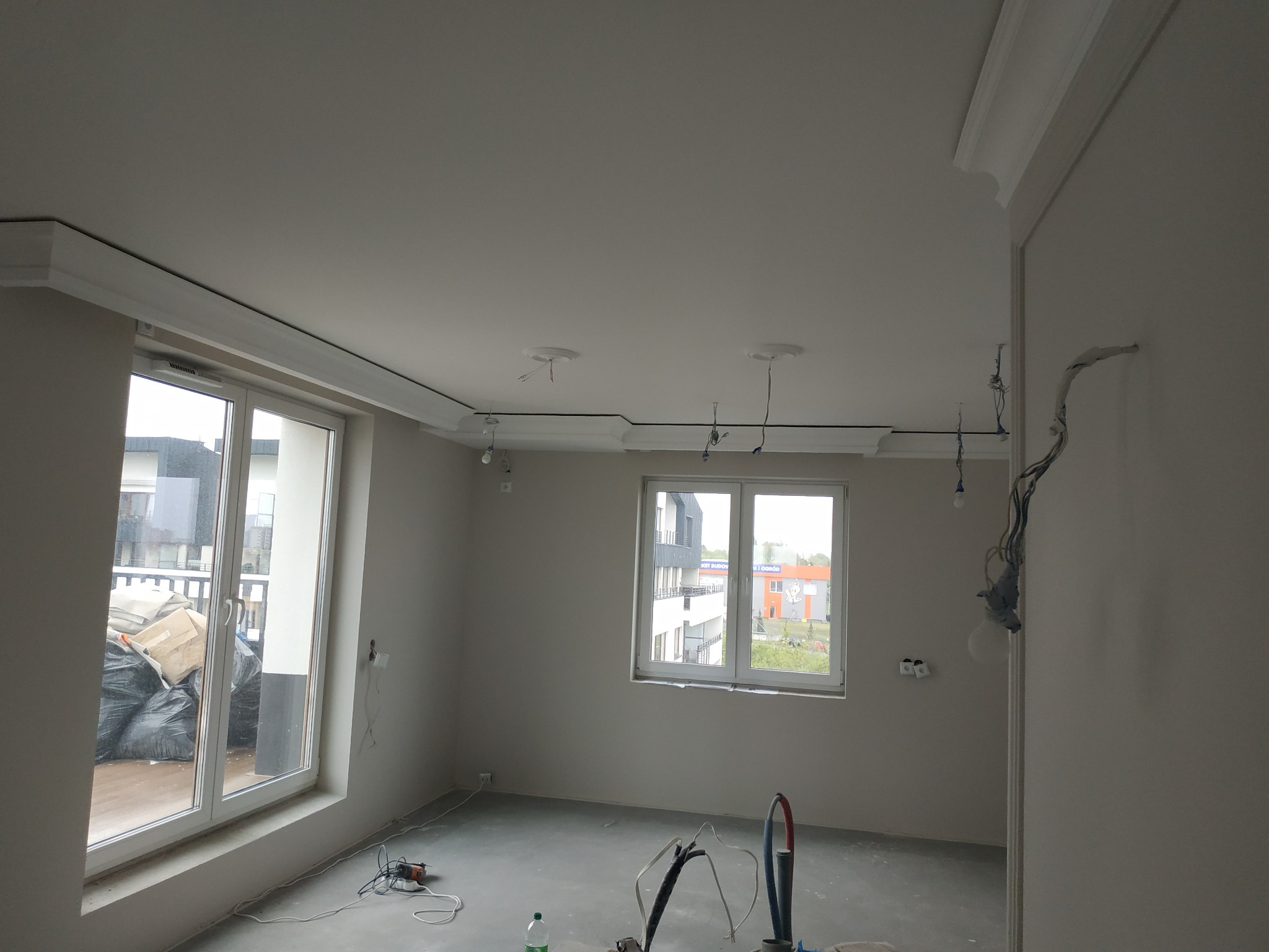Pokój po szpachlowaniu i wstępnym malowaniu, widoczne kable od oświetlenia, listwy dekoracyjne pod sufitem, okno balkonowe z workami gruzu na zewnątrz, przygotowany podkład podłogowy.