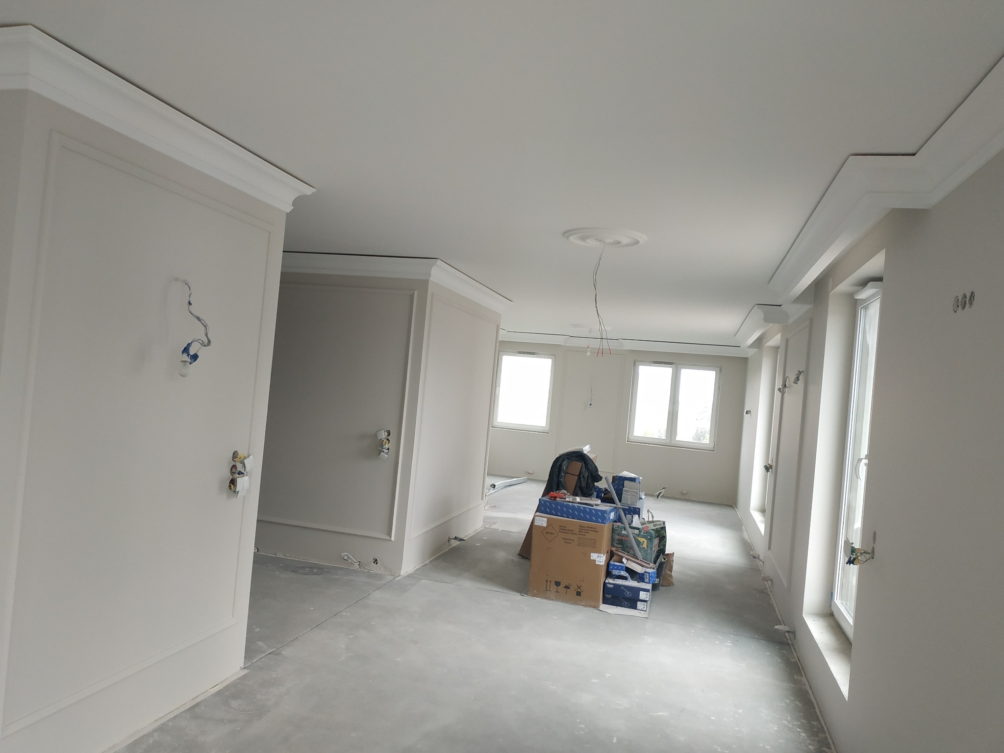Wnętrze w trakcie remontu: ściany z dekoracyjnymi listwami i gładzią, surowy beton na podłodze, widoczne puszki elektryczne i materiały budowlane w kartonach.