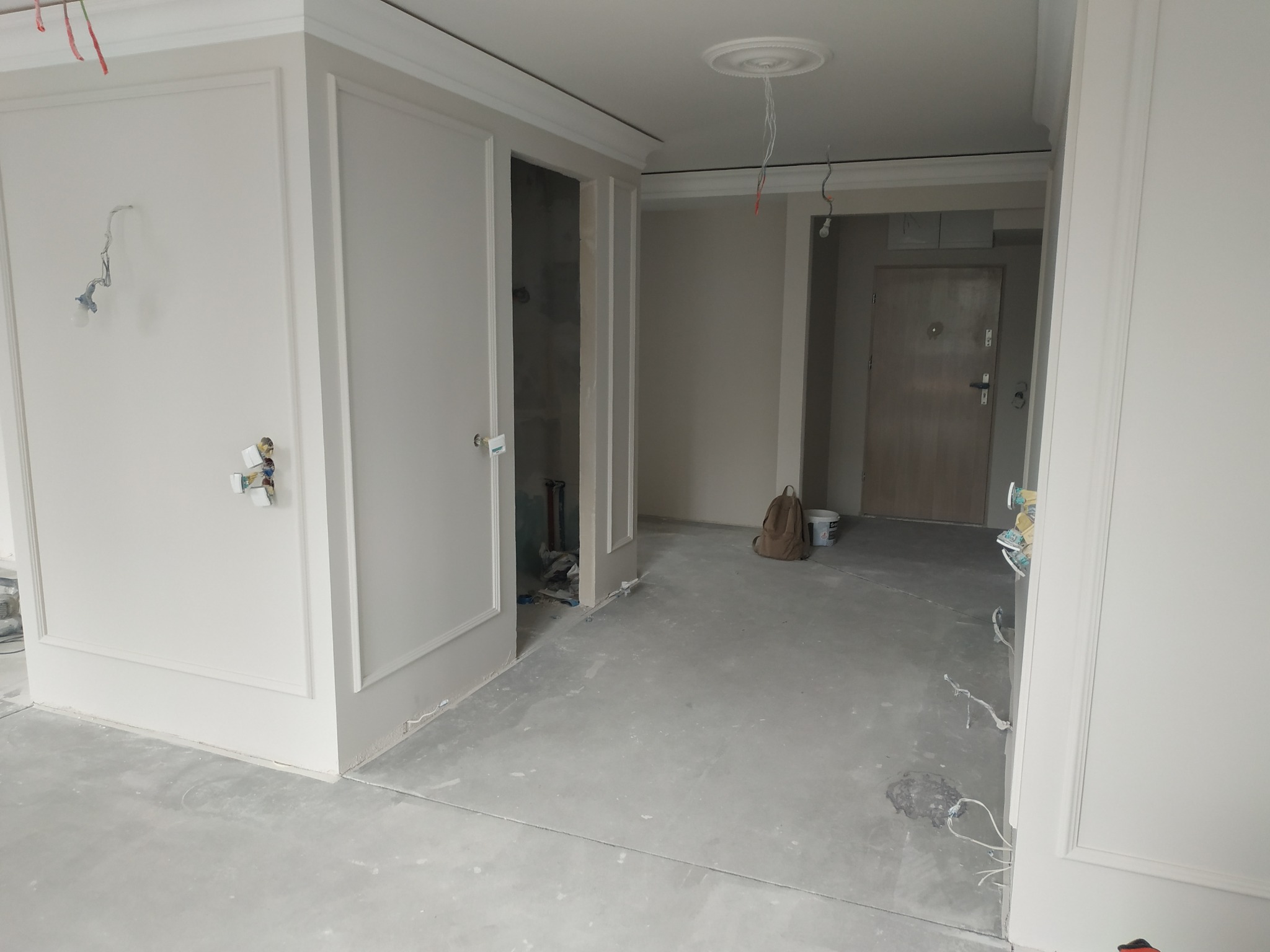 Wnętrze w trakcie remontu: białe ściany z dekoracyjnymi listwami, surowy beton na podłodze, widoczne kable elektryczne, plecak i wiadro w tle, sugerujące przerwę w pracach wykończeniowych.