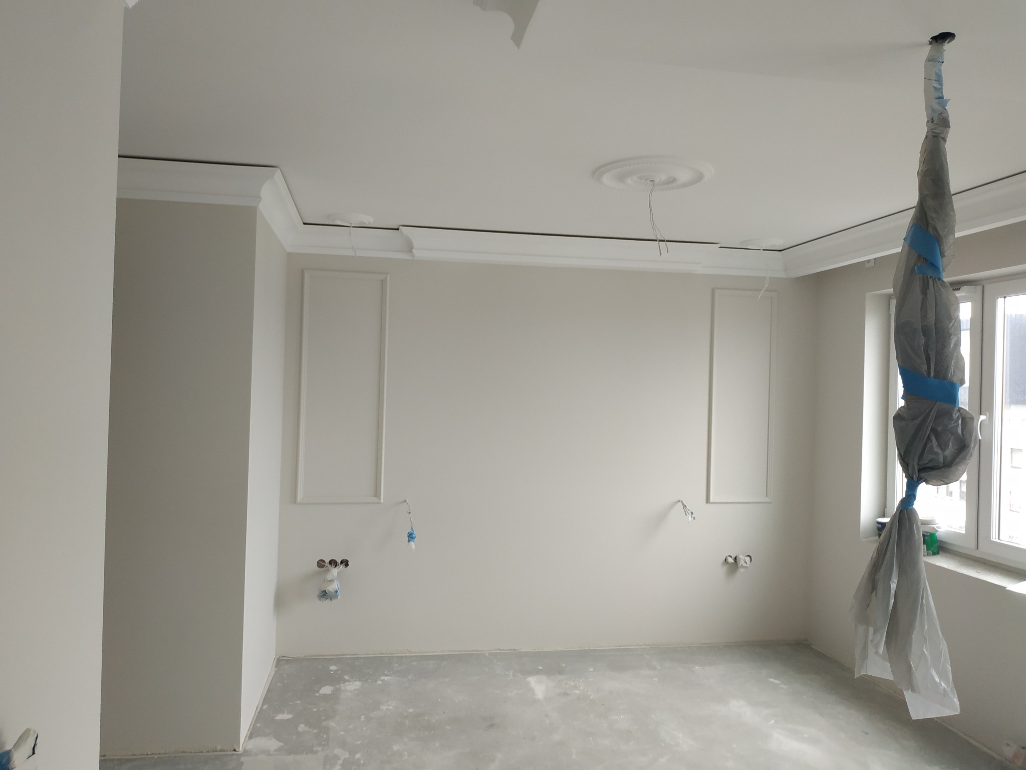 Pokój w trakcie remontu: ściany z dekoracyjnymi panelami i listwami, zabezpieczone instalacje elektryczne, podłoga z wylewką, okno osłonięte folią malarską.