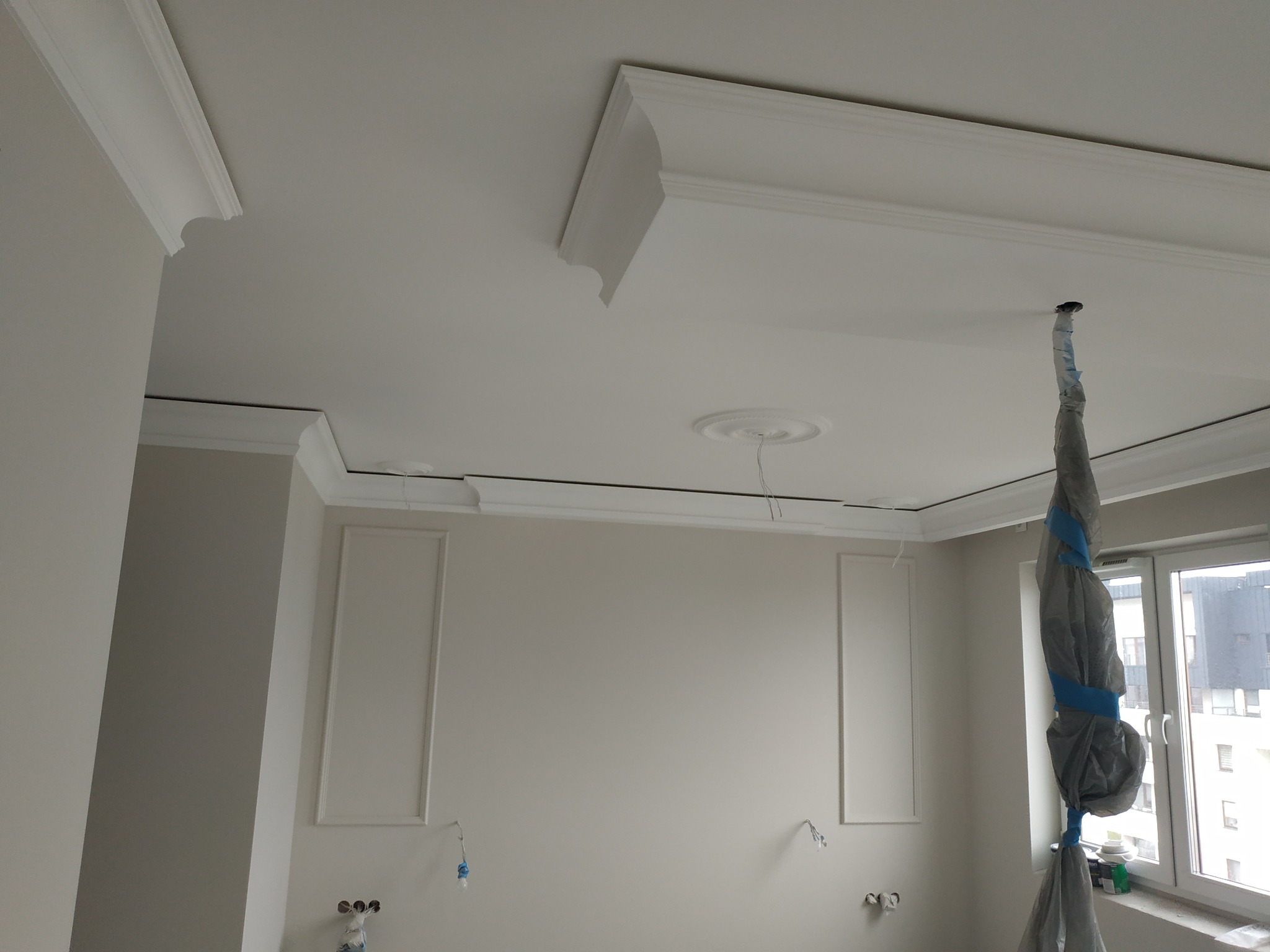 Jasne wnętrze w trakcie wykańczania: ściany z dekoracyjnymi panelami, białe listwy przysufitowe z ornamentami, zabezpieczona folią lampa wisząca, okno z widokiem na budynki.